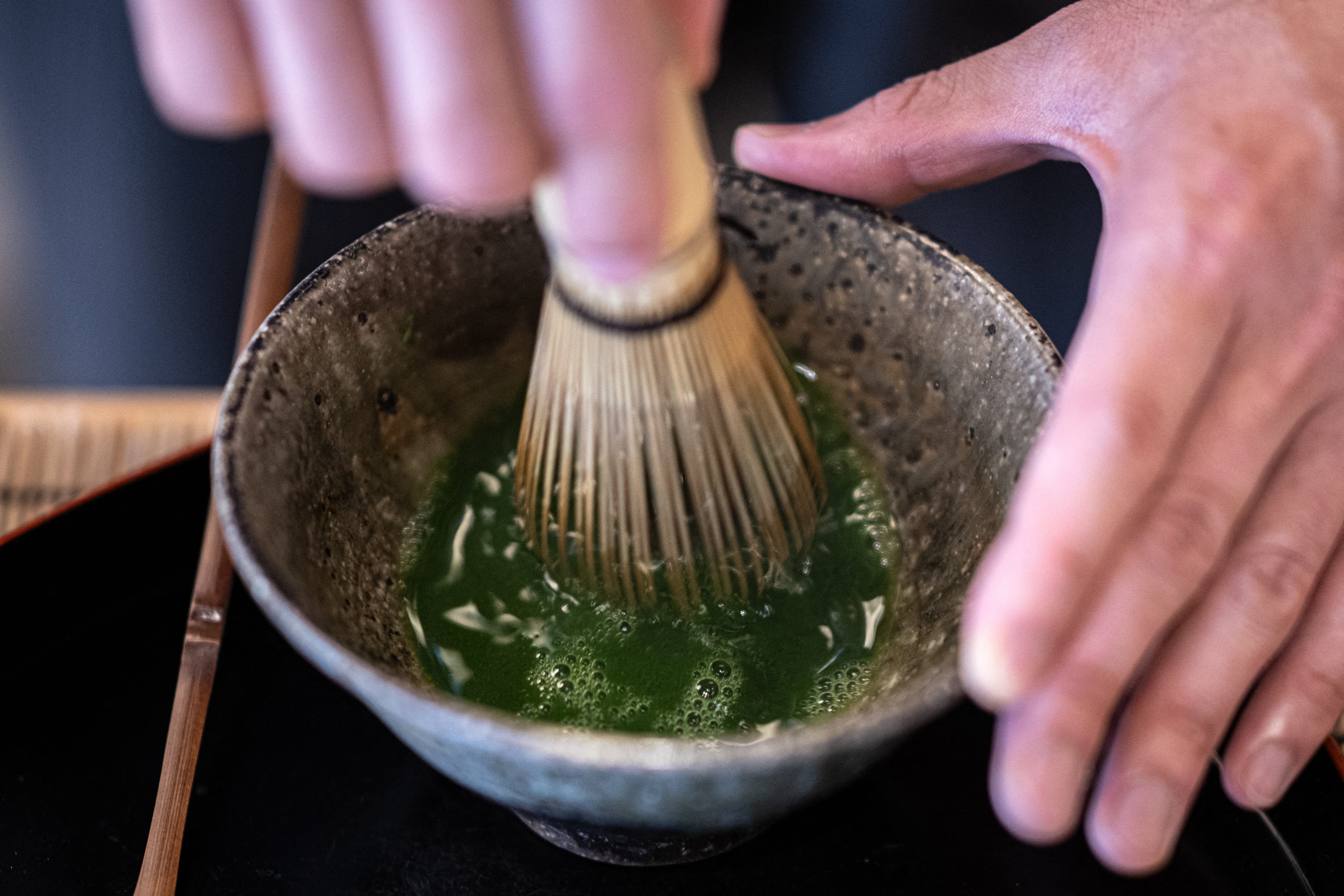 A person holds a whisk while mixing a matcha powder and water in a cermaic bowl.