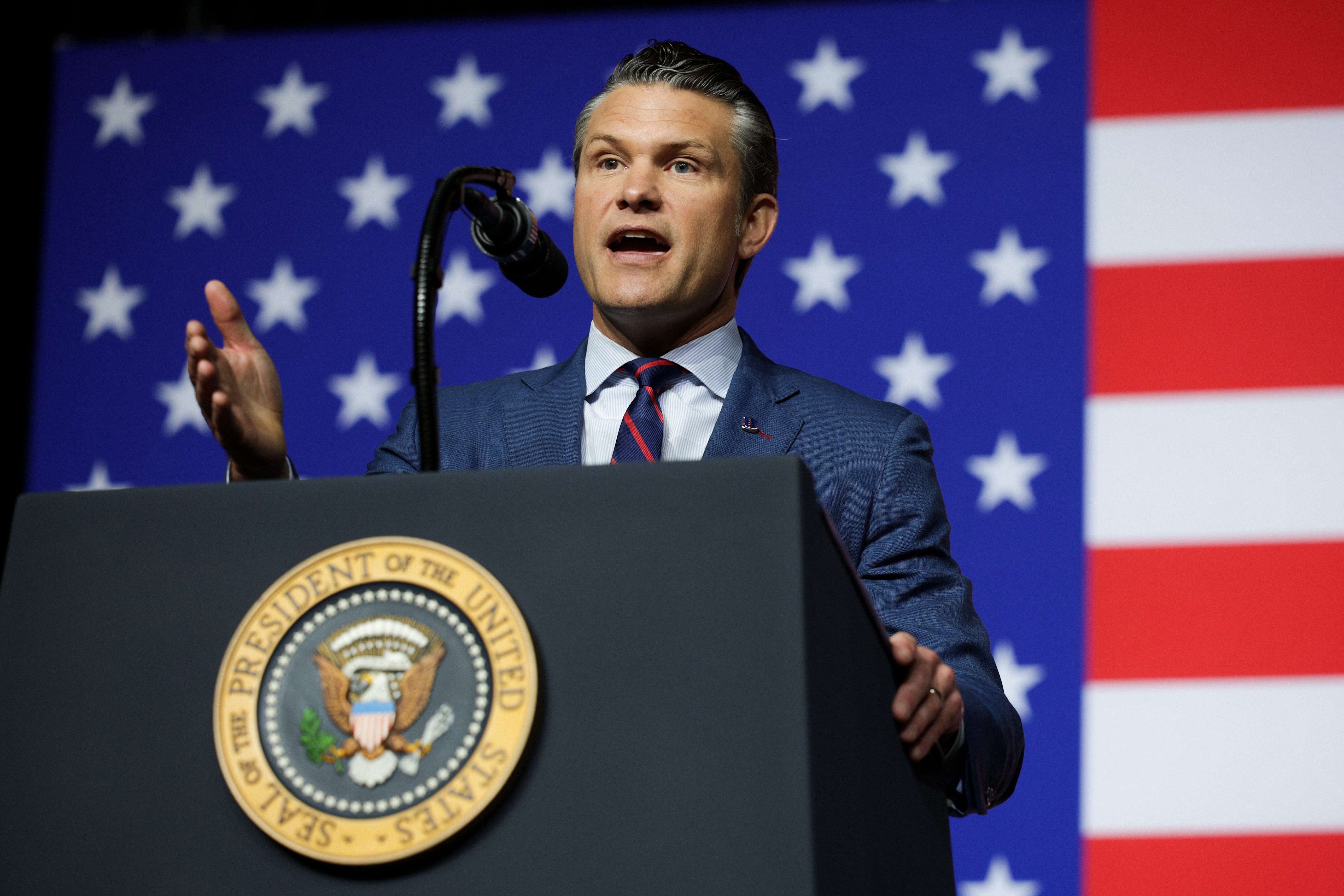 US Secretary of War Pete Hegseth speaking at a podium with the American flag in the background