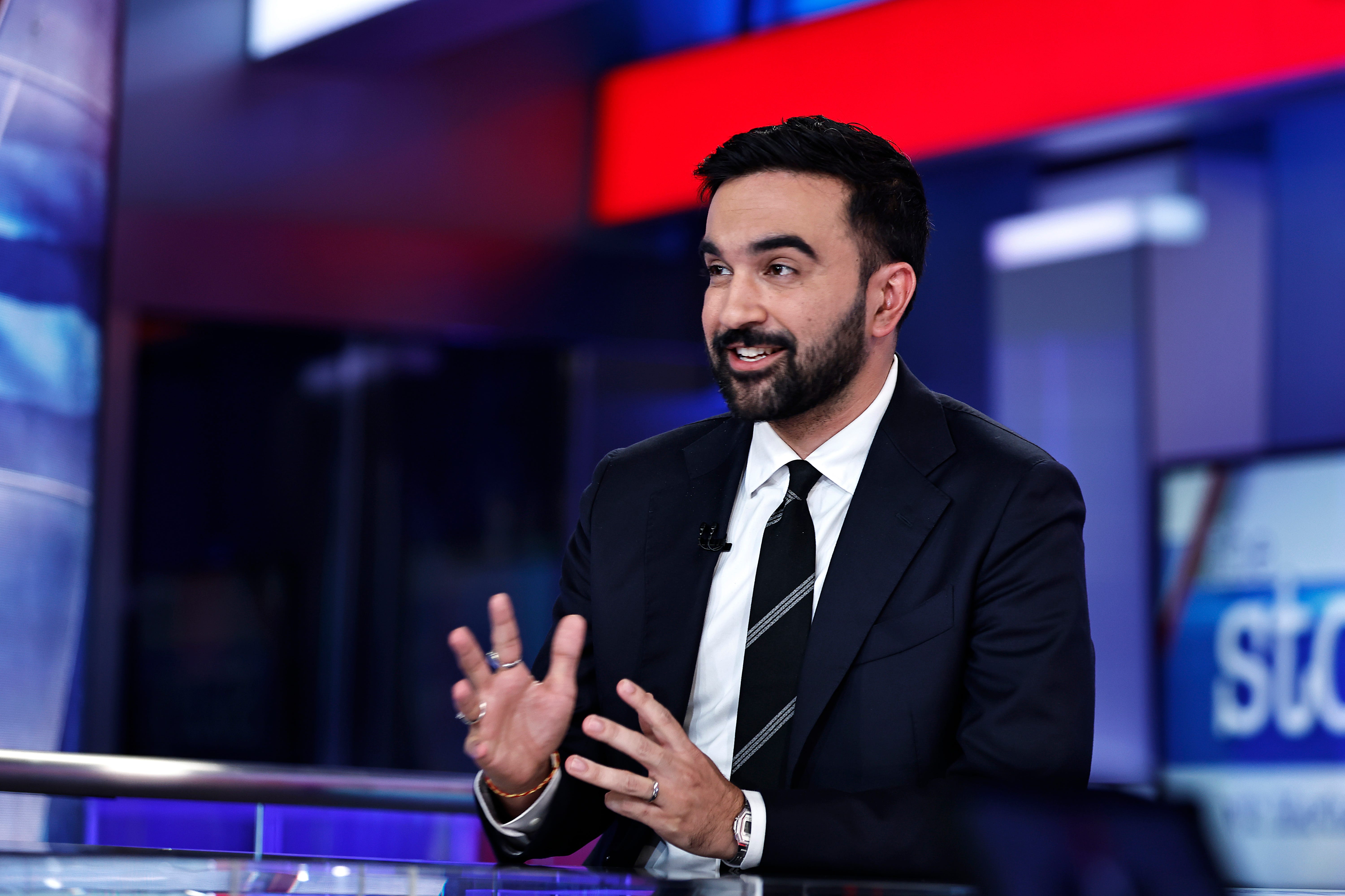 Zohran Mamdani in a dark suit and white shirt sitting looking animated.