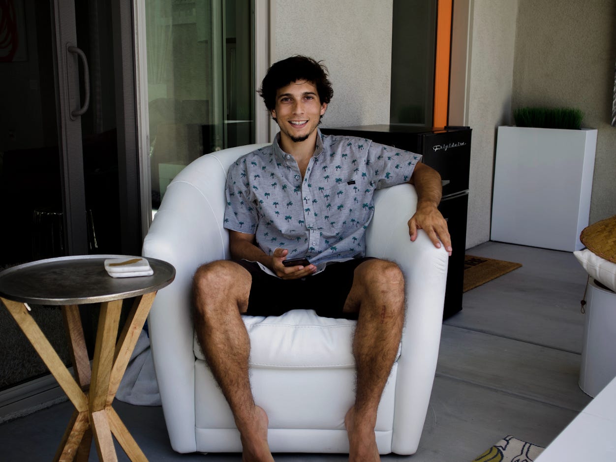 Man with dark hair sits in white chair and poses in a short sleeve printed shirt and shorts
