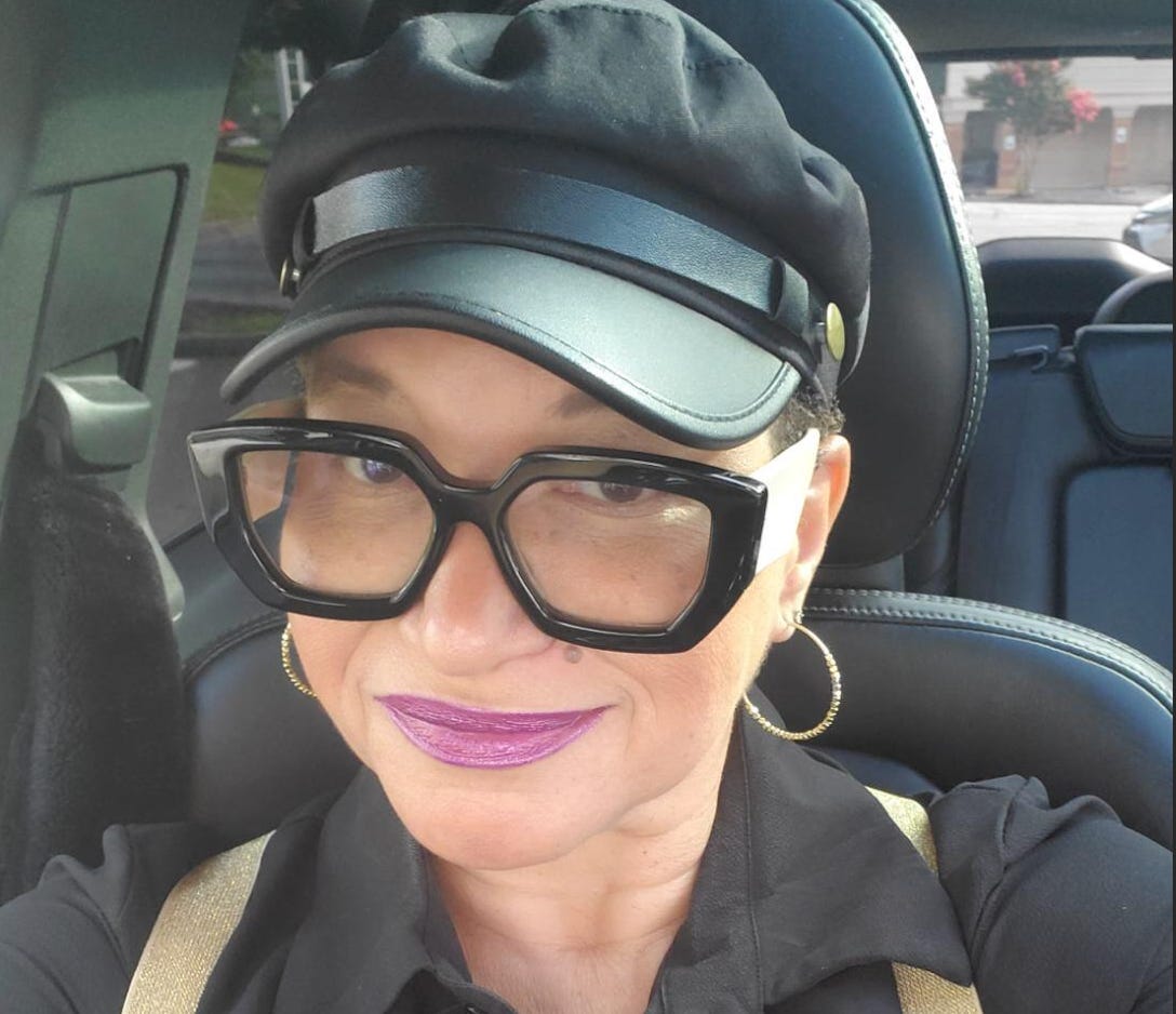 Anja Holthoff, an Uber driver in Atlanta, looks at the camera while wearing a black paperboy cap, black-rimmed glasses, and a black blouse with suspenders.