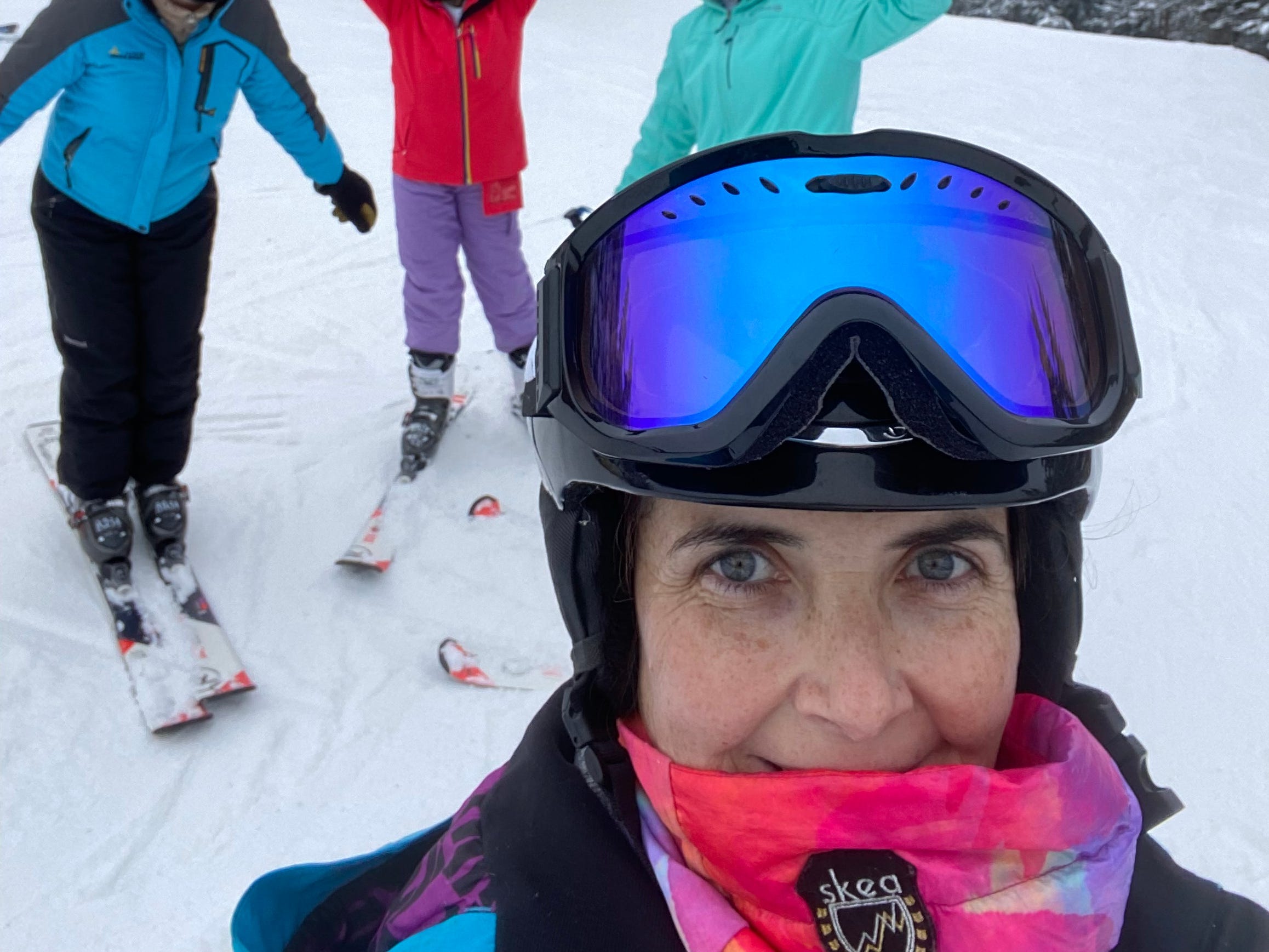 A woman is taking a selfie on a ski slope with three people behind her.