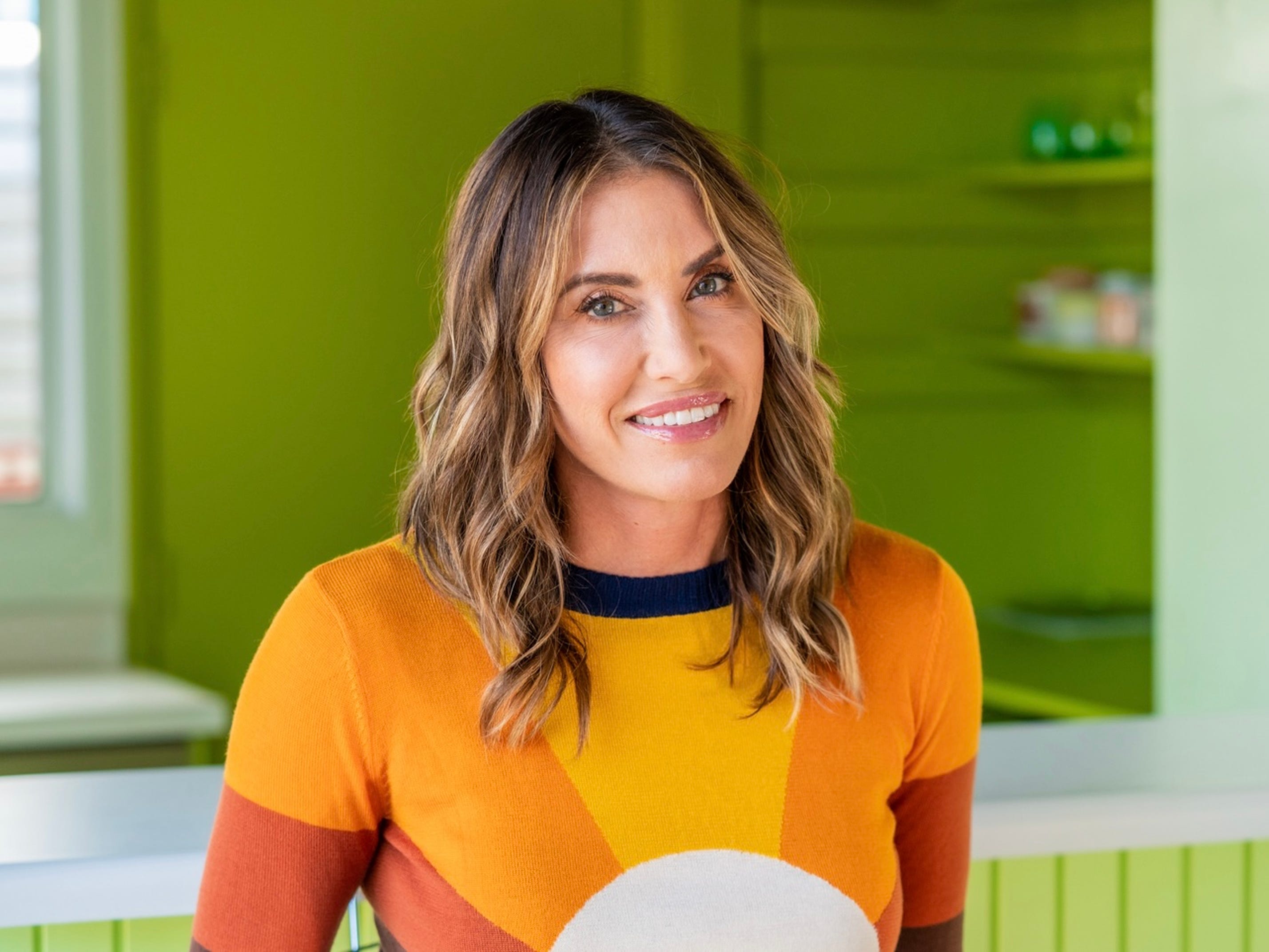 Wende Zomnir wearing a yellow and orange sweater and smiling in a bright green room.