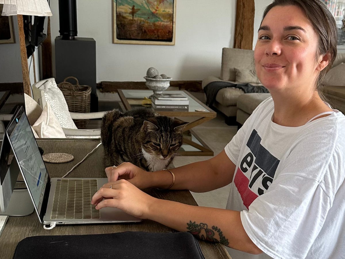 The author working at her laptop while her cat sits on a desk next to her.