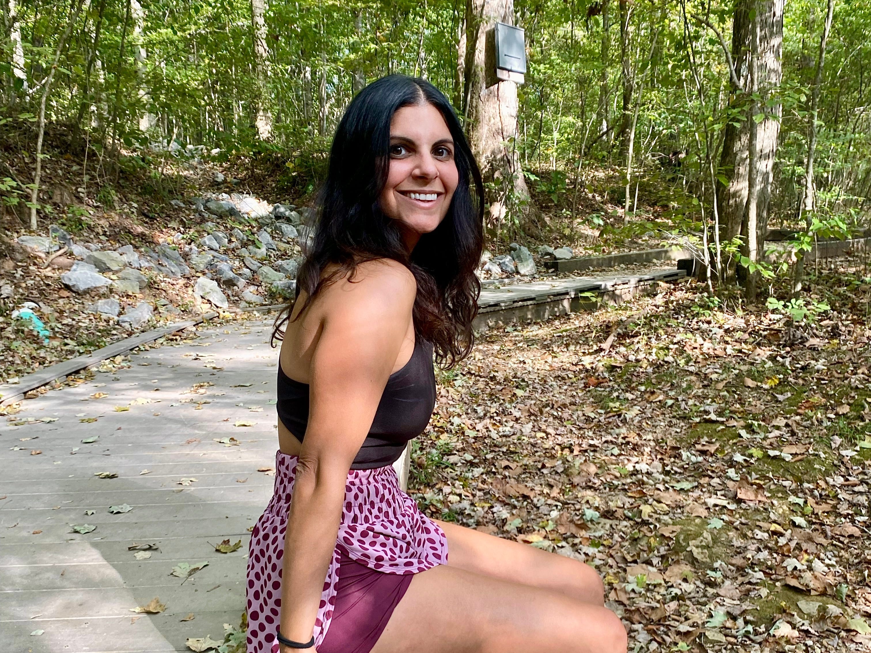 Woman sitting on a wooden railing in a forest, smiling at the camera, wearing a sports top, shorts, and slip-on shoes.