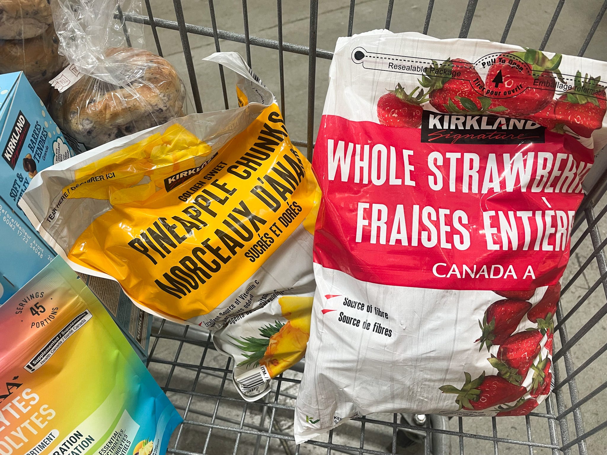 A close-up of items in the author's cart, including frozen strawberries and pineapples, blueberry bagels, granola bars, and Organika electrolyte powder sachets.