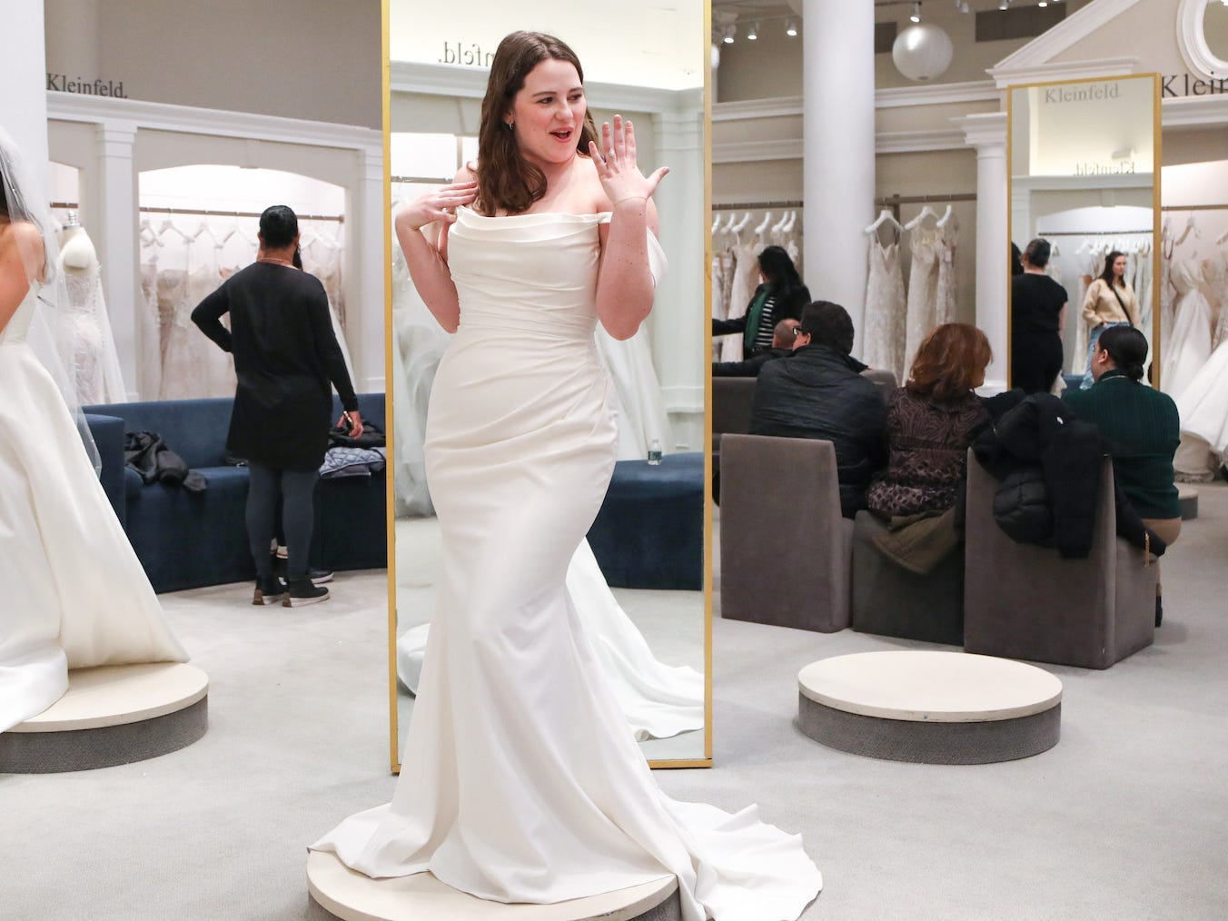 A woman holds up her ring finger while she wears a wedding dress in a bridal boutique.