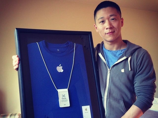 A person stands indoors holding a framed display containing a blue Apple Store T-shirt, an employee badge, and a business card. The person is wearing a gray zip-up hoodie over a blue shirt and is smiling at the camera.