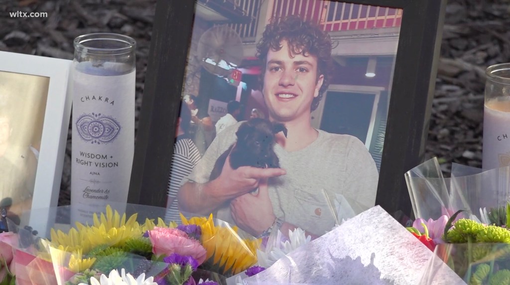A framed photo of a smiling young man holding a puppy, surrounded by bouquets of flowers and candles.