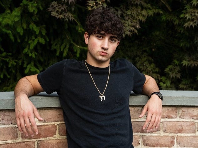 A young man leans against a brick wall outdoors, wearing a black T-shirt and a gold necklace.