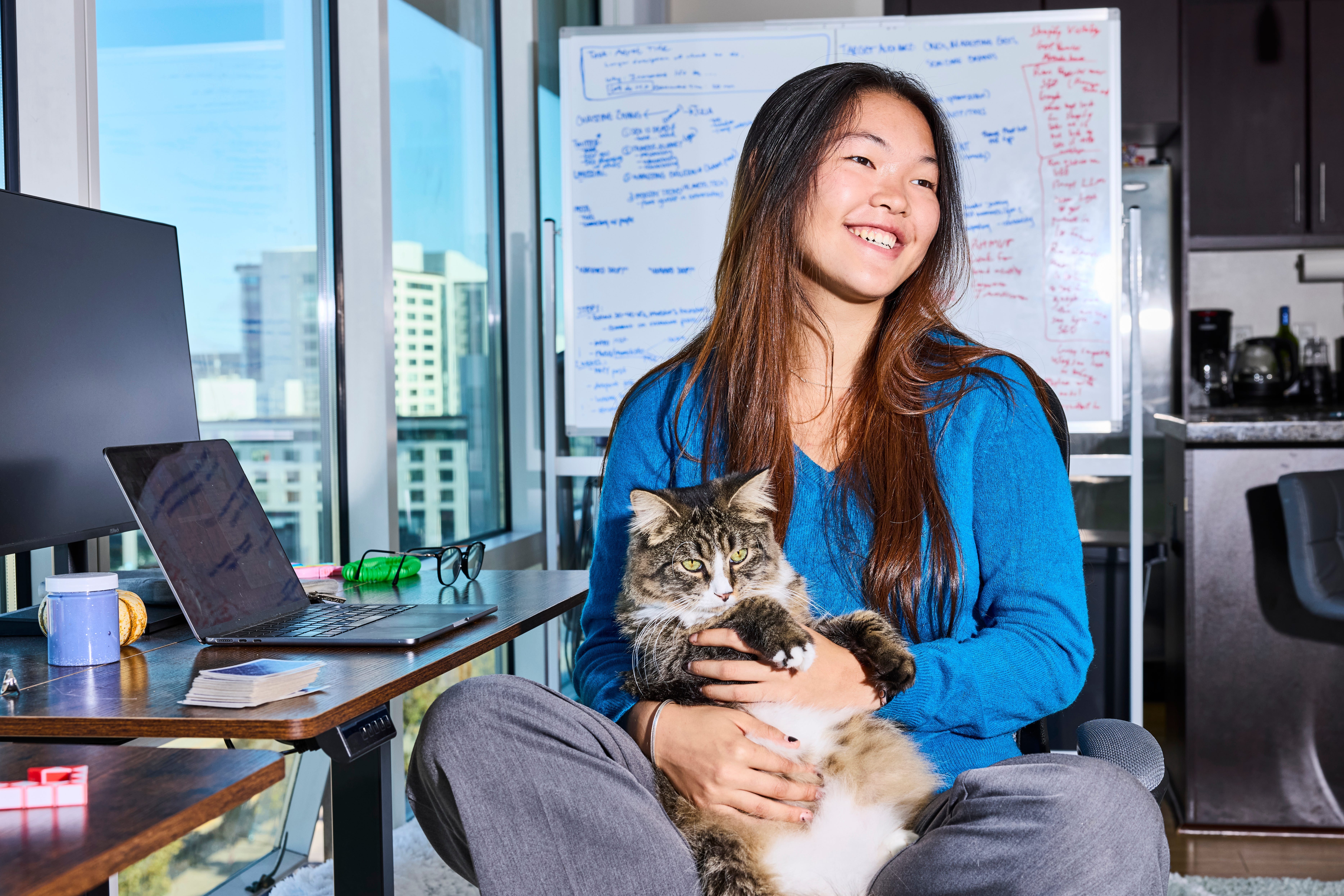A girl holding her cat sits in front of a desk and is smiling off to the side