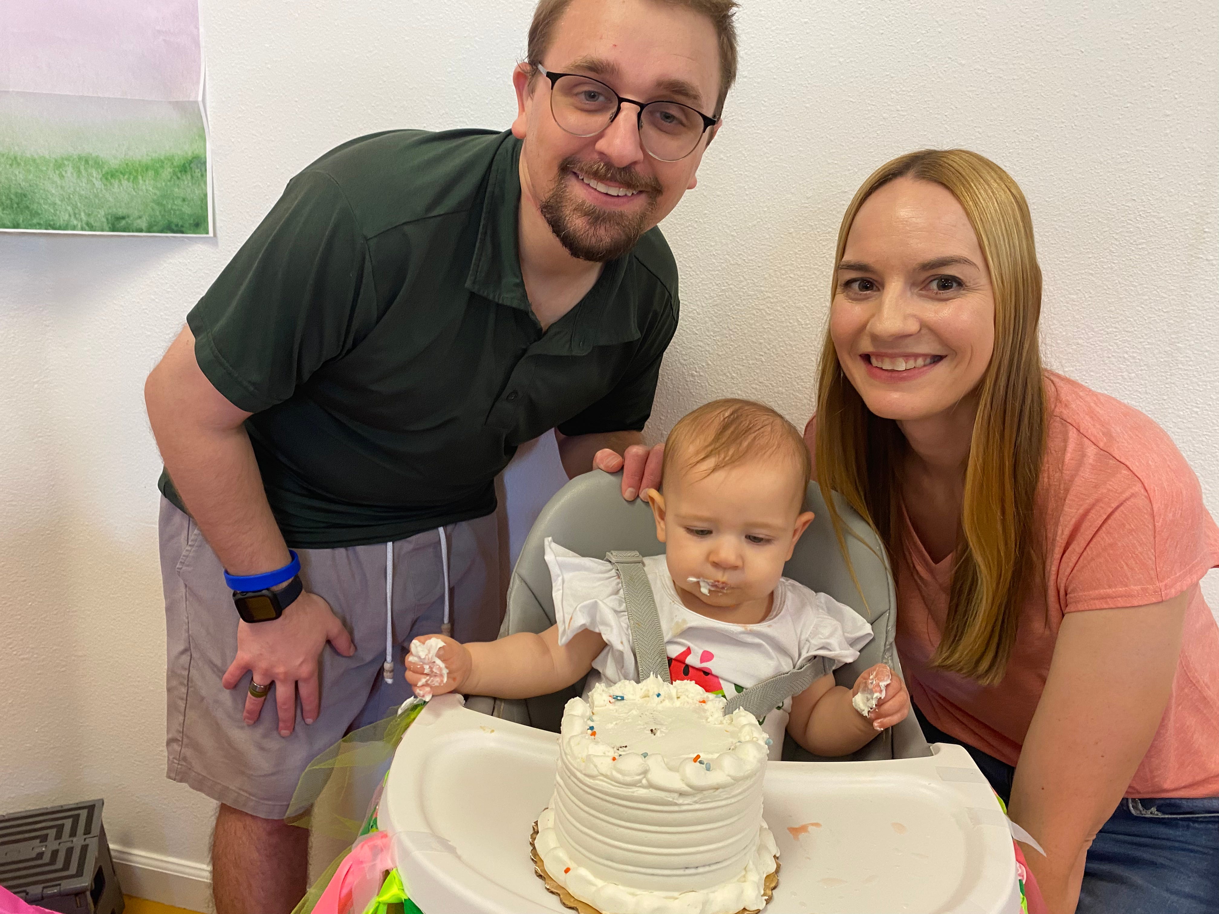Man and woman lean over toddler in highchair with birthday cake