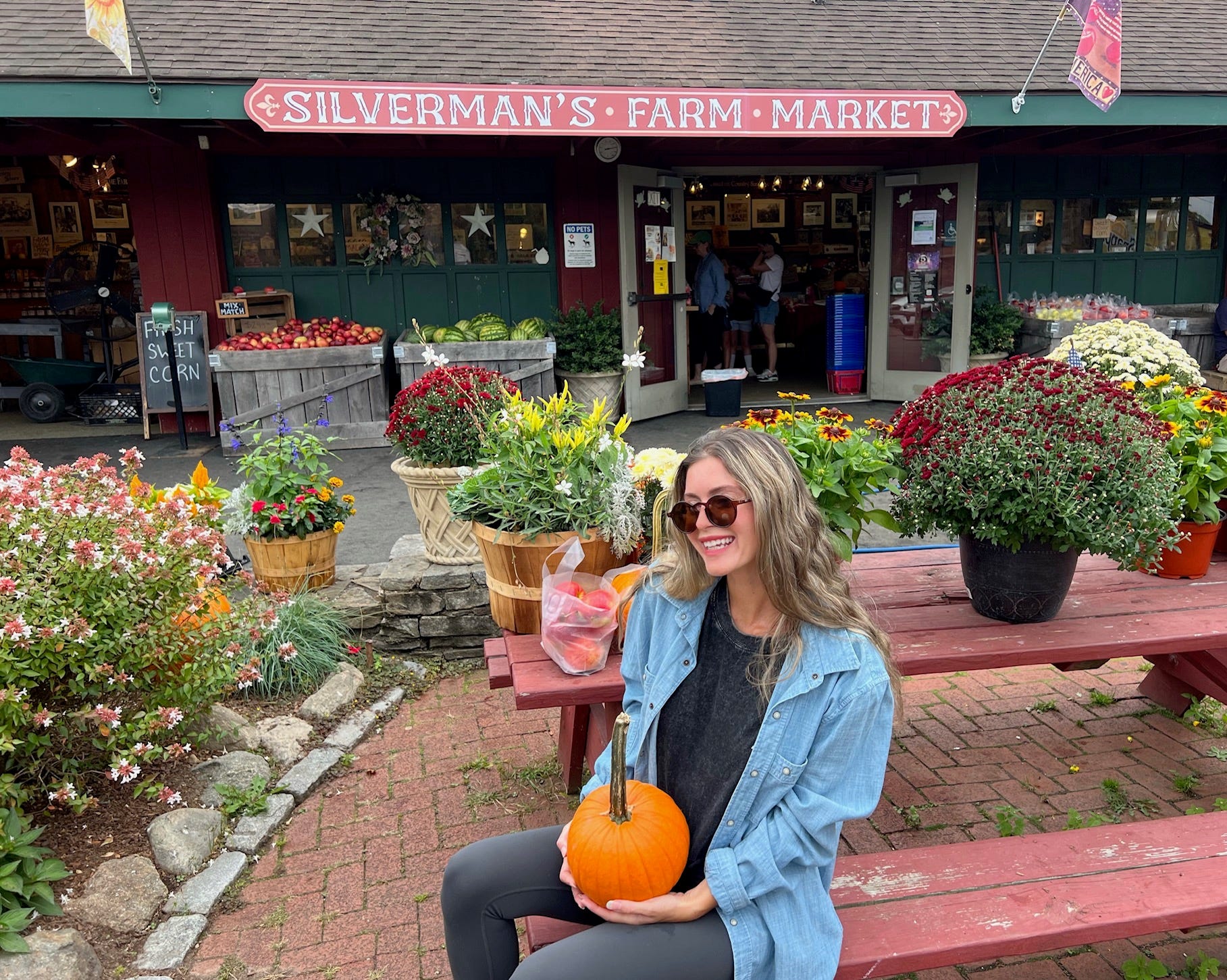 Author Alexa Mellardo holding pumpkin on picnic bench in front of Silverman's Farm Market
