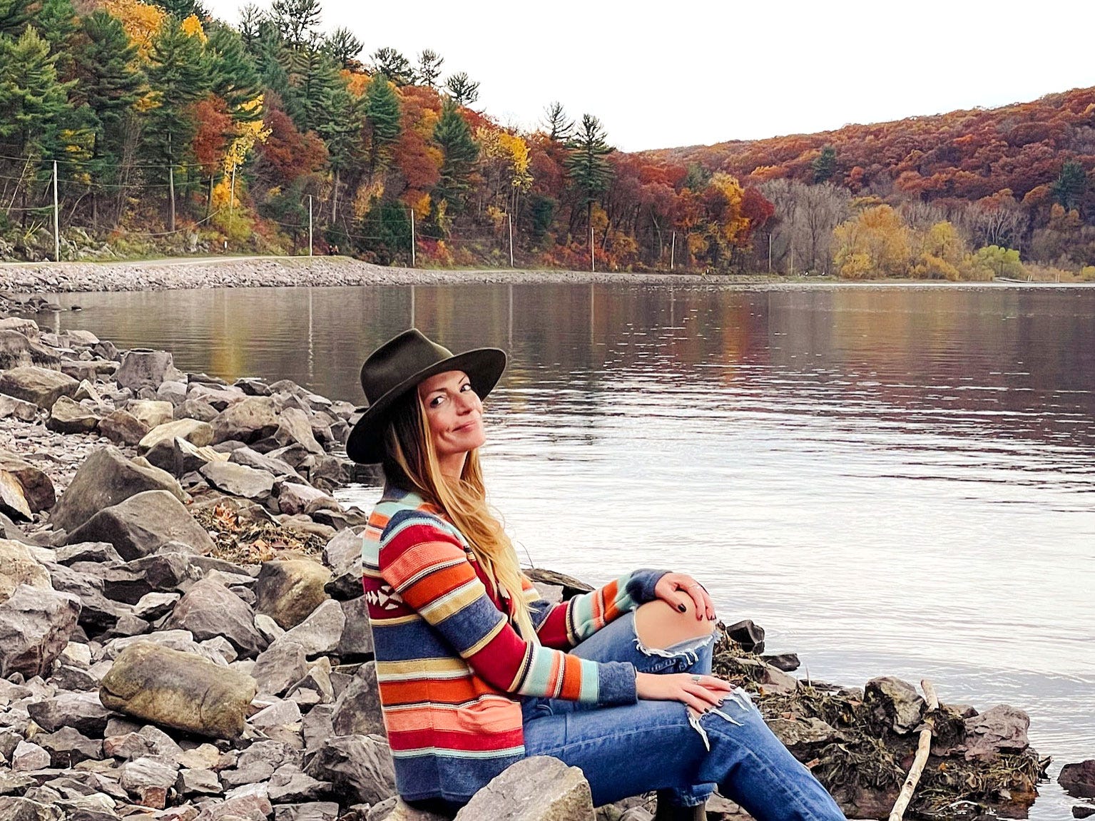 Emily sits on the bank of a lake, with fall foliage in the background.