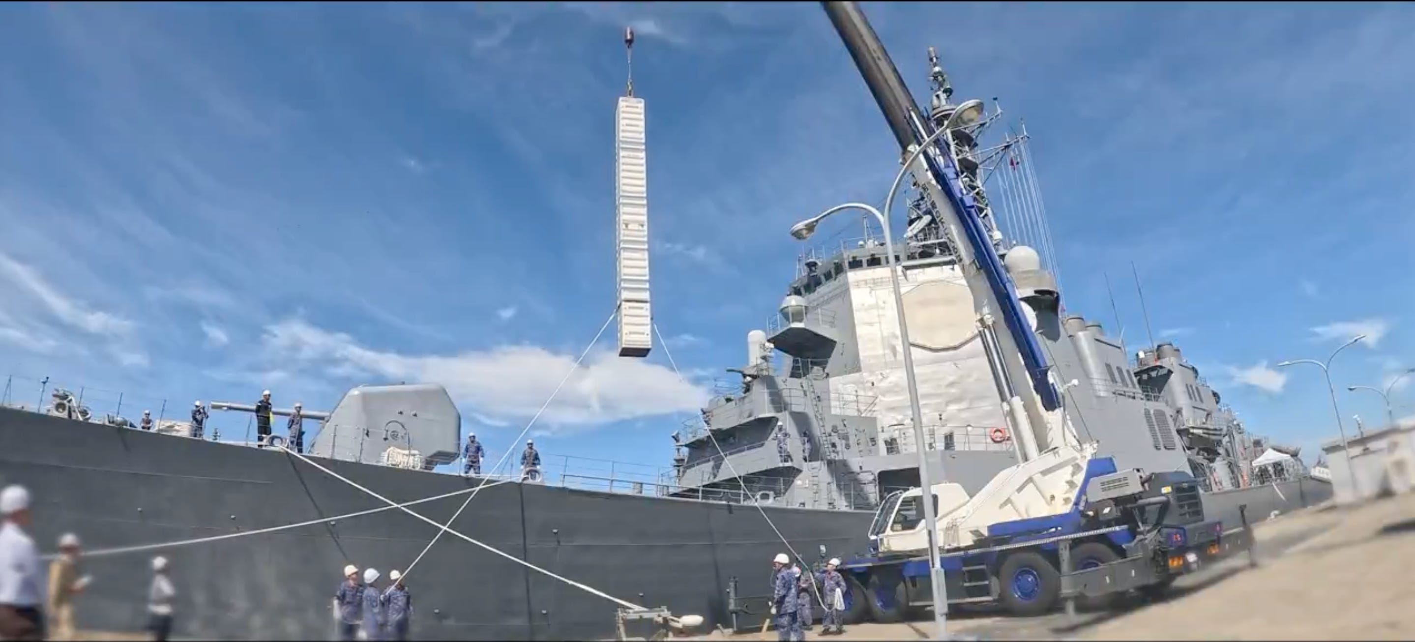 A Japanese warship is docked with a large crane putting a large white cylinder on top of the ship. The sky is blue and cloudy in the background.