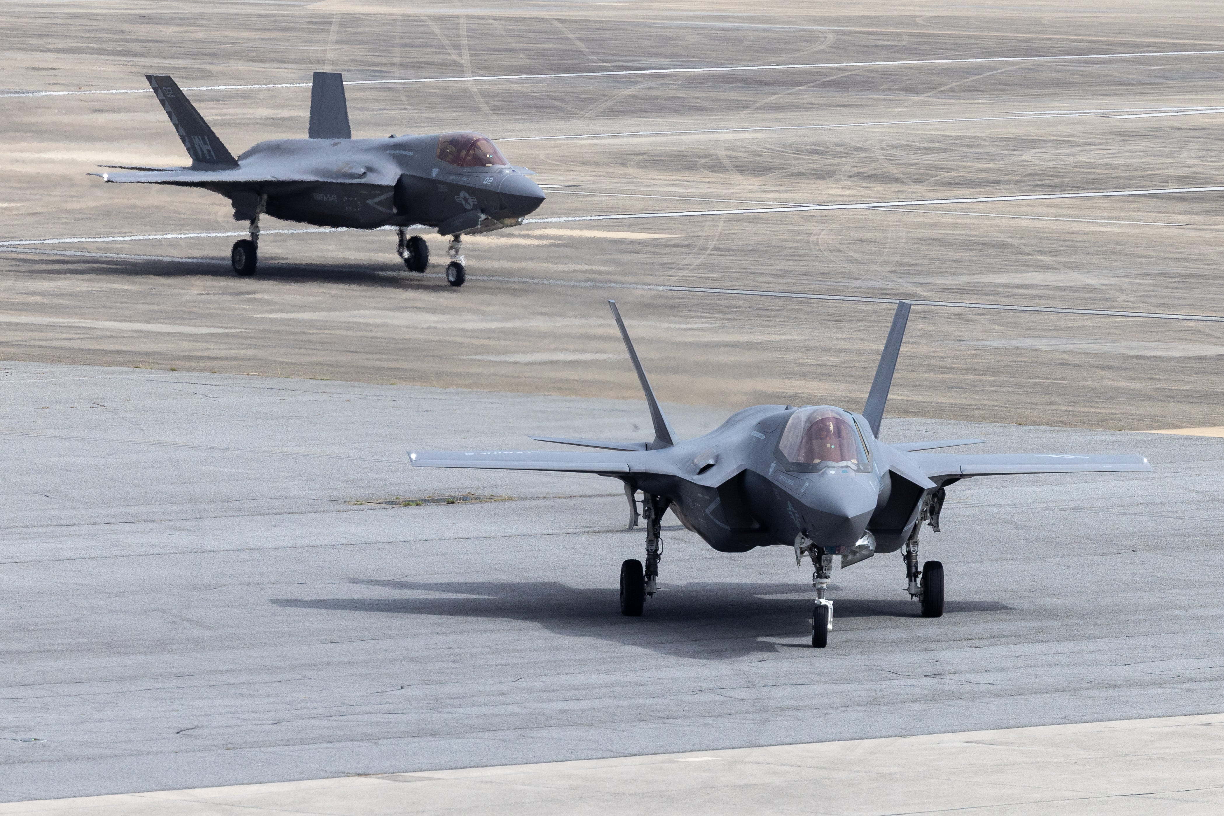 Two Marine Corps F-35B Lightning II stealth tigers sit on a tarmac. There is a line of grass in the background.