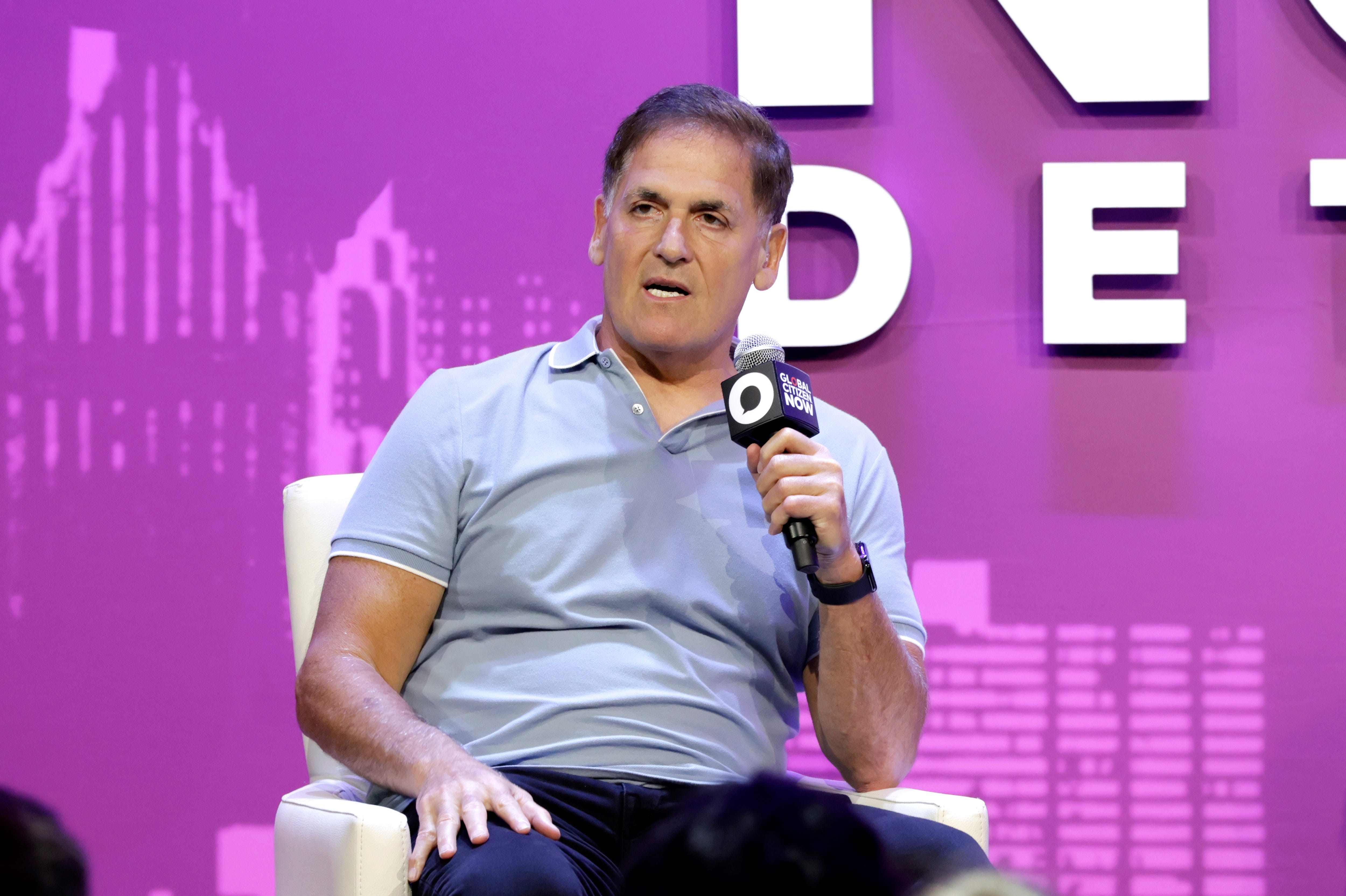 Mark Cuban is wearing a gray polo T-shirt and holding a microphone with his left hand.