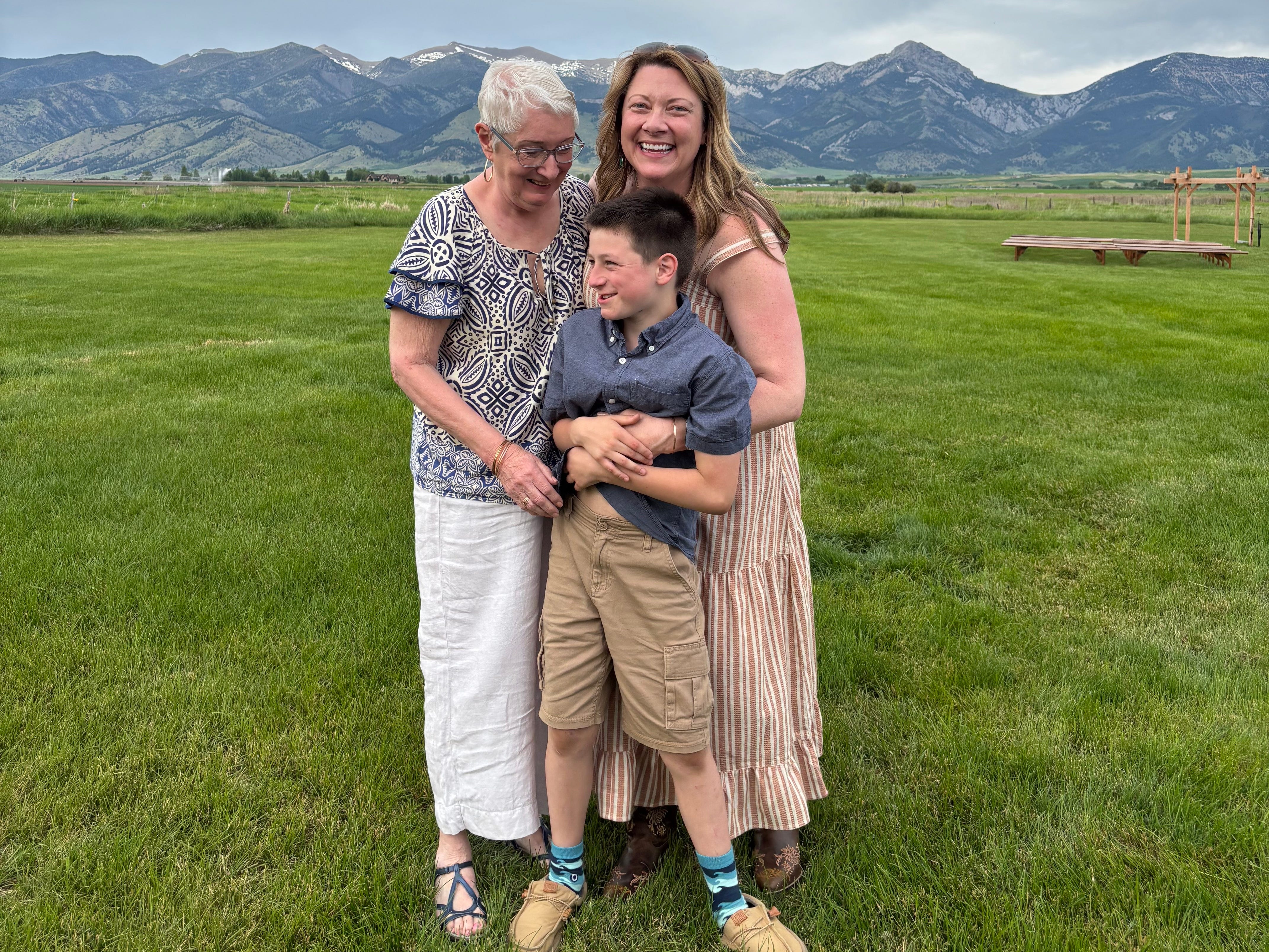 A grandmother, her adult daughter and grandson pose in front of a mountain range.