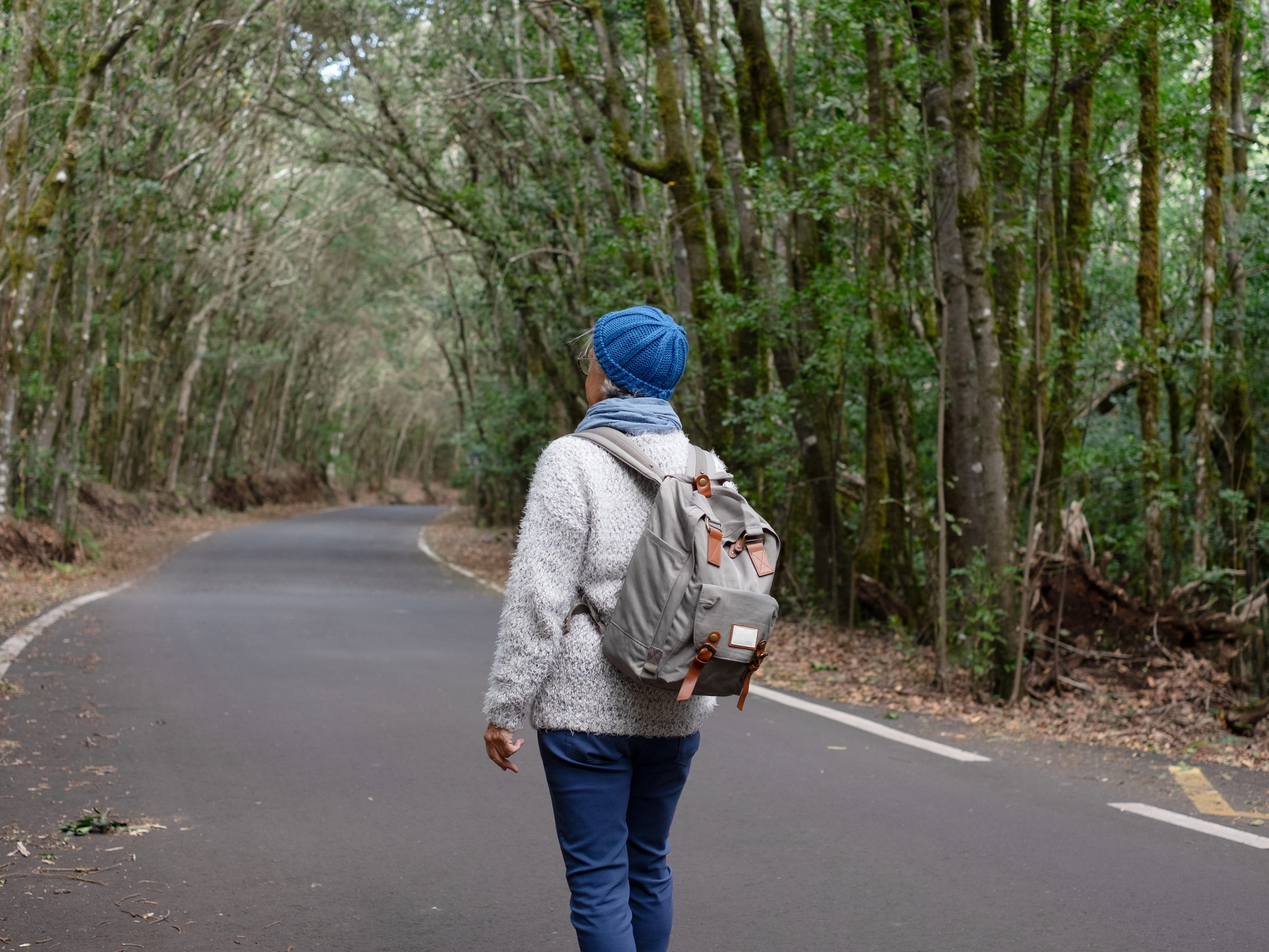 A woman wearing a sweater, a backpack, and a knitted blue beanie walking down a wooded path. Her back is to the camera.