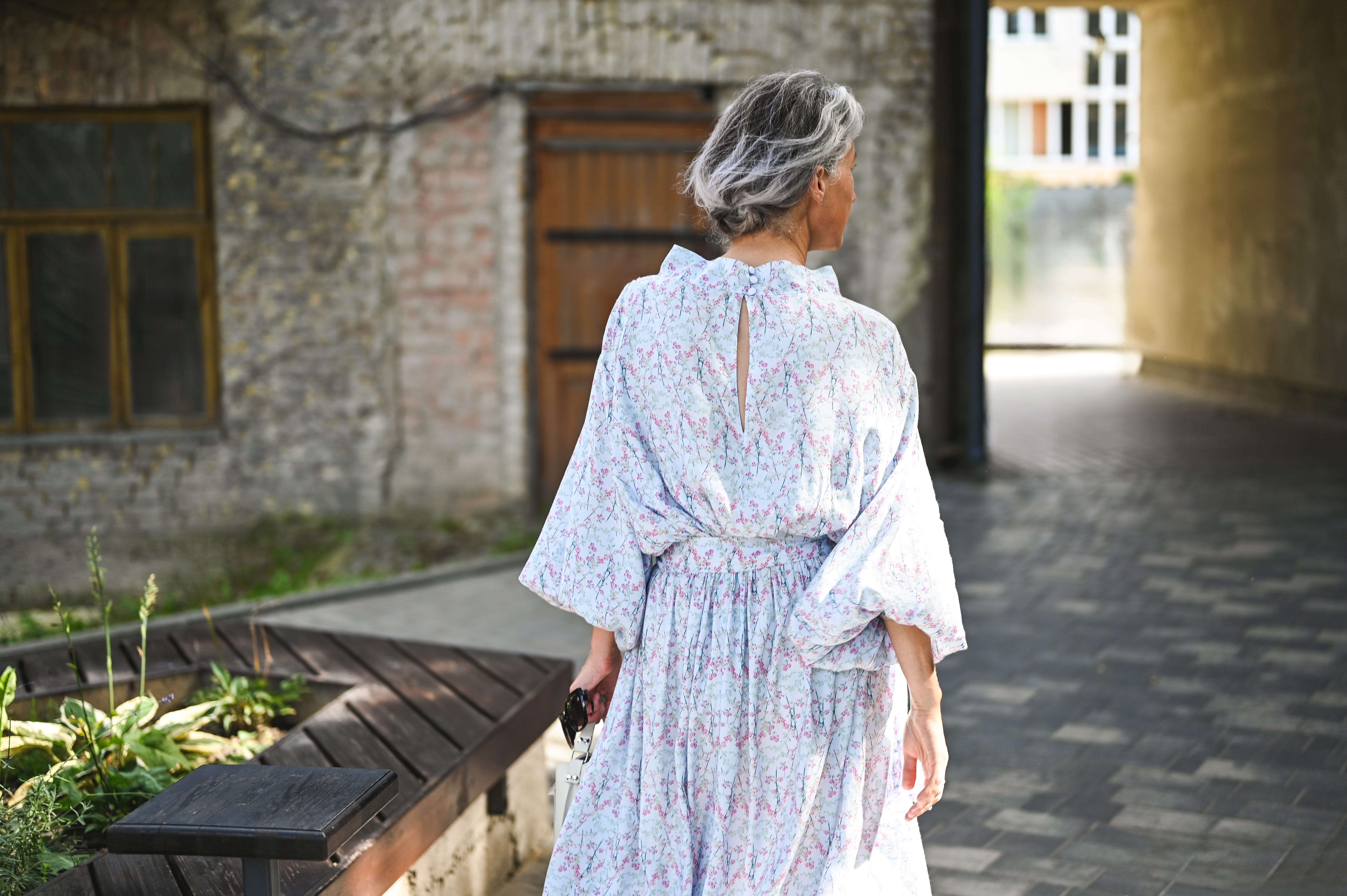 Woman with gray hair, flowy dress facing away from camera in courtyard