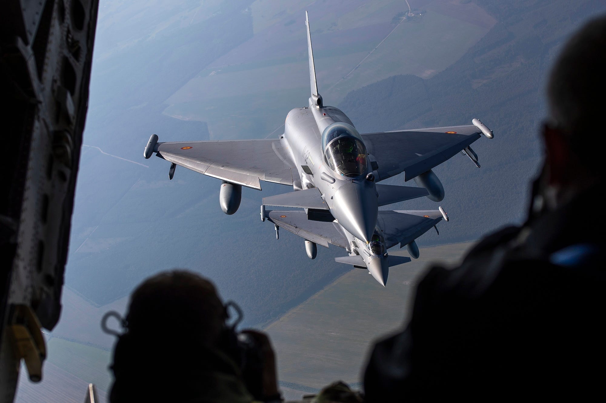 A grey fighter jet in the air pictured from the inside of another aircraft flying above, with the silhouette of some people in front of the camera visable in the foreground