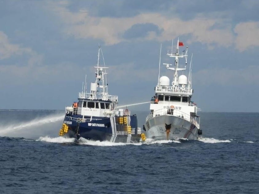 A white Chinese coast guard ship sprays a water cannon on a blue Philippine Coast Guard vessel in dark blue waters with a cloudy blue sky in the background.