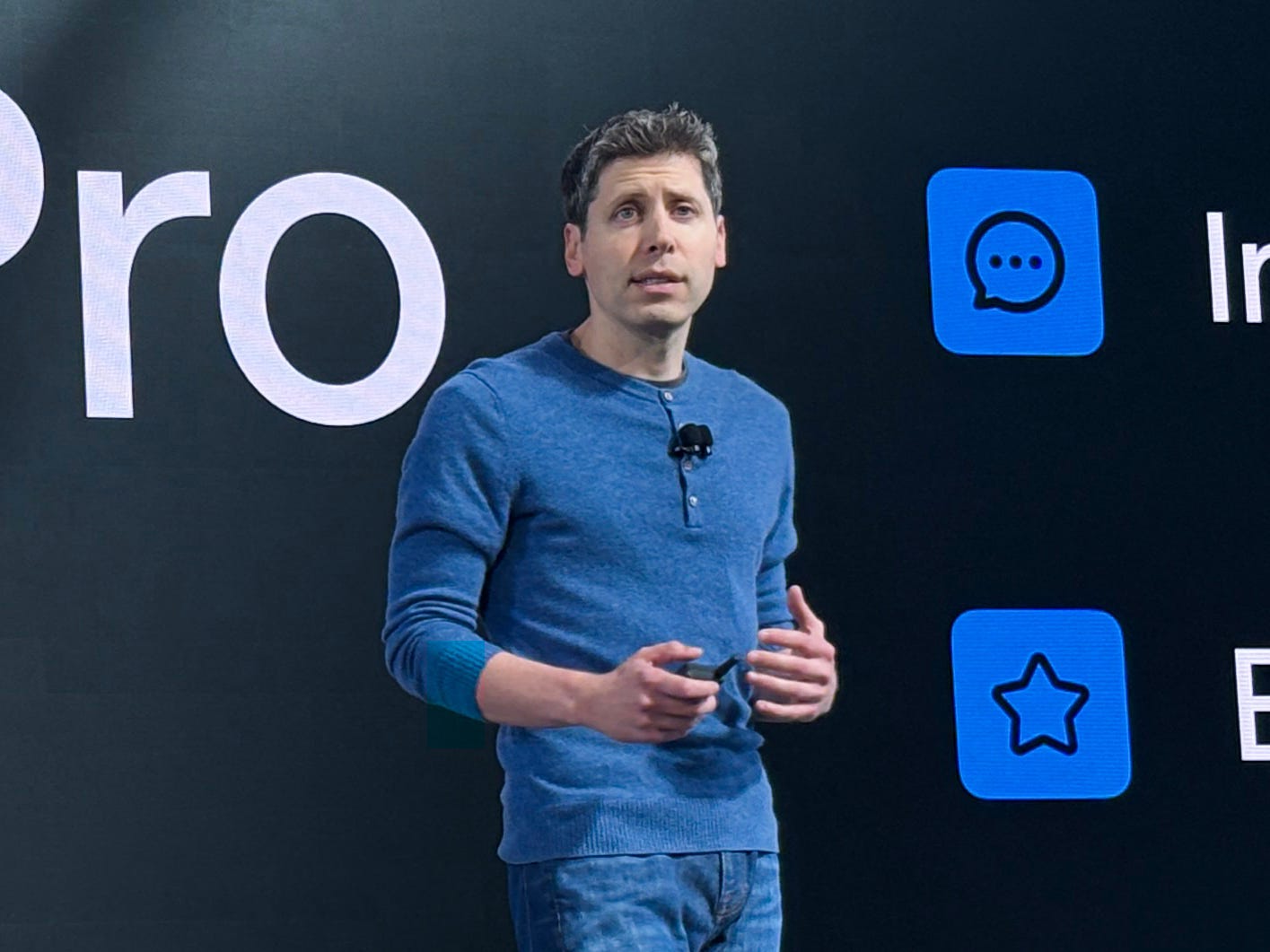 Sam Altman standing on stage at DevDay, wearing a blue shirt and jeans.