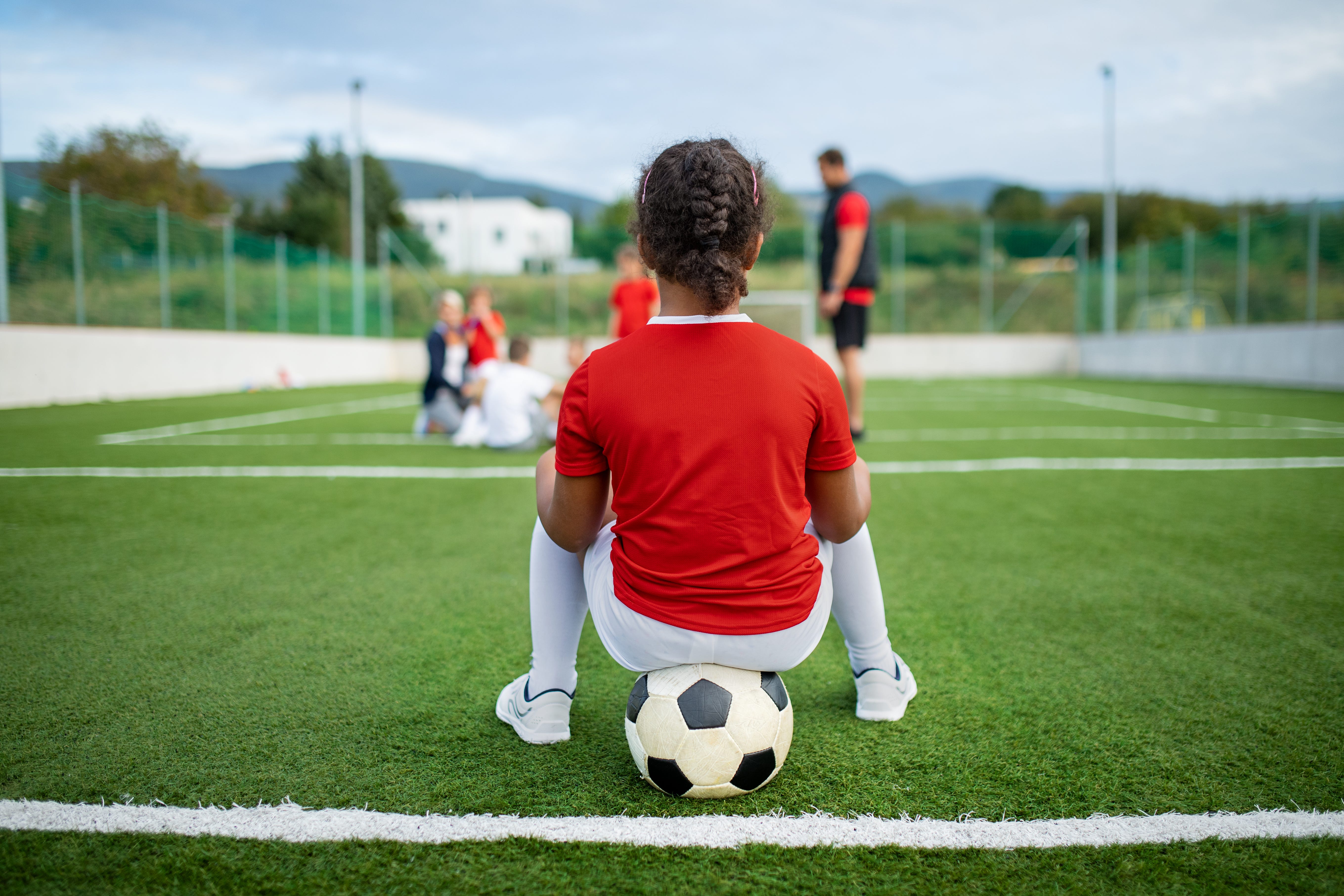 A girl wearing a red shirt and white socks balances on a soccer ball on the side of a field.