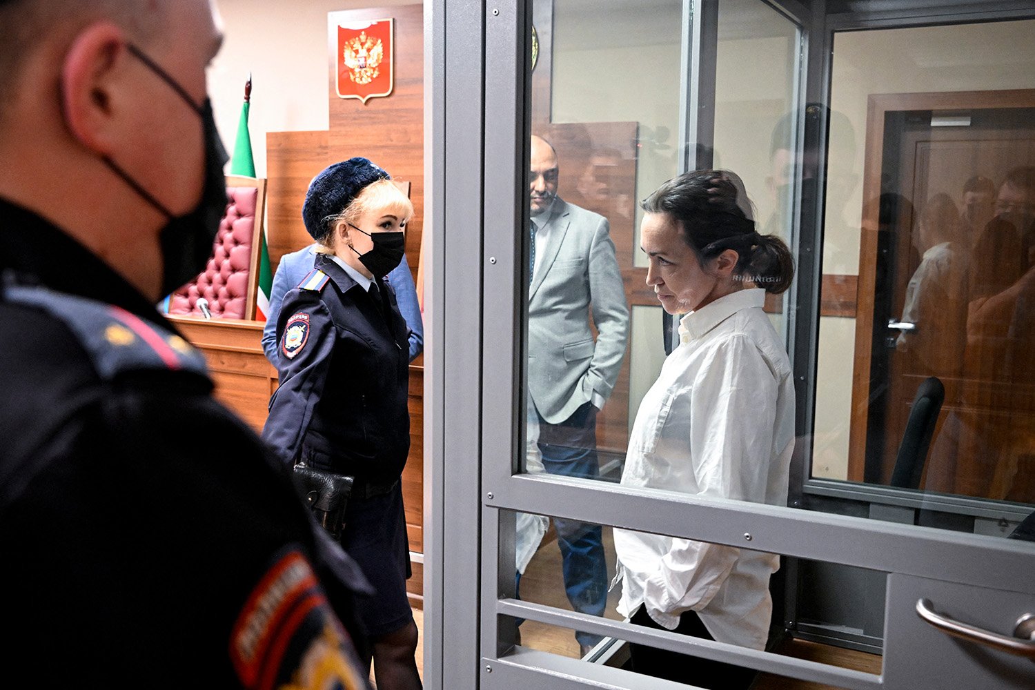 A woman stands in a holding cell, dressed in a white button down, and looks through the glass at three Russian police officers.