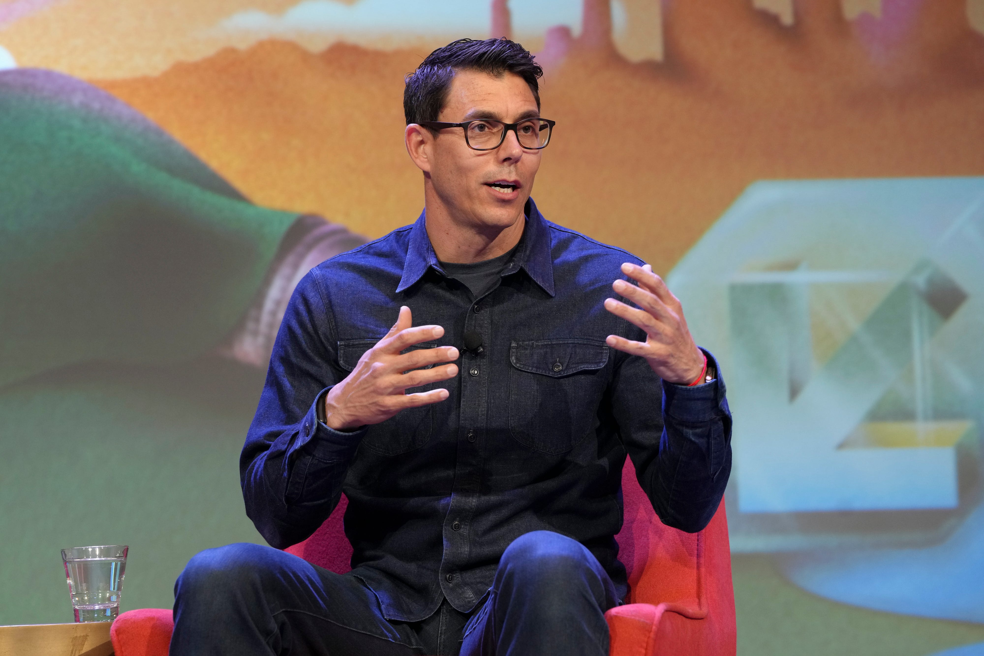 Rivian CEO RJ Scaringe is wearing a black shirt and sitting on a red sofa.