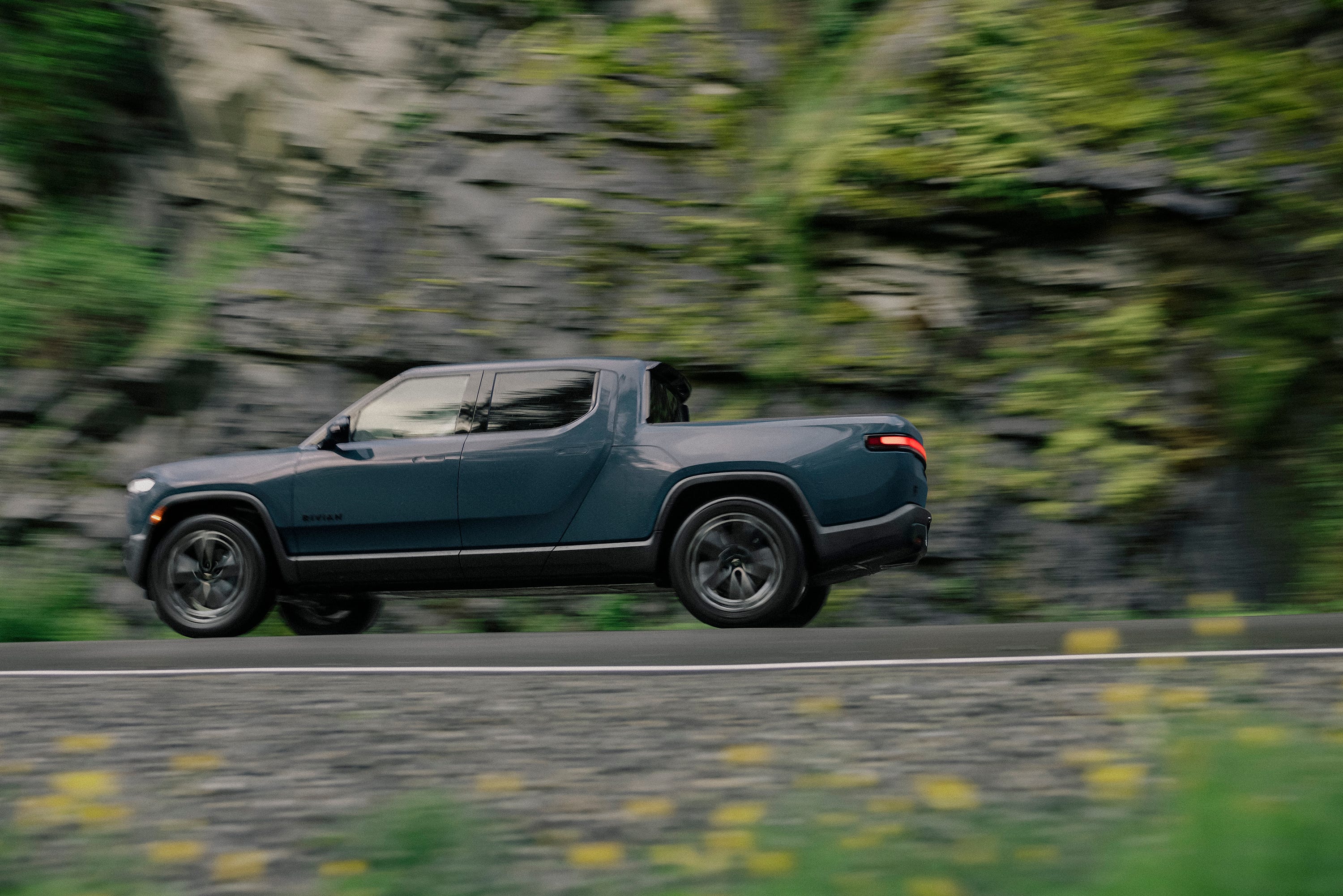 A blue Rivian R1T pick up truck driving down a mountain road.