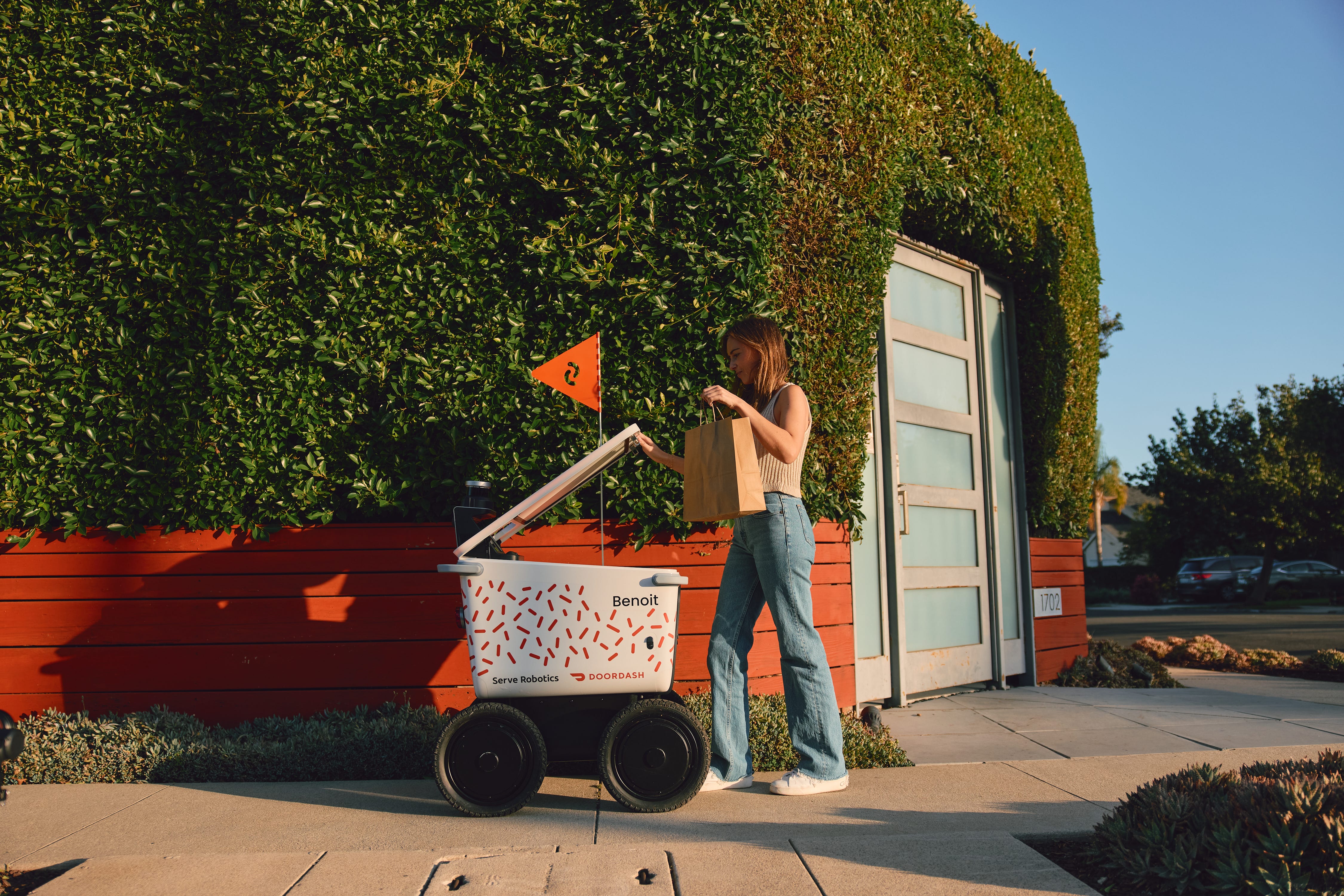 A woman holds a paper shopping bag above one of Serve's four-wheeled delivery robots, which has a white body with a red, triangular flag above it.