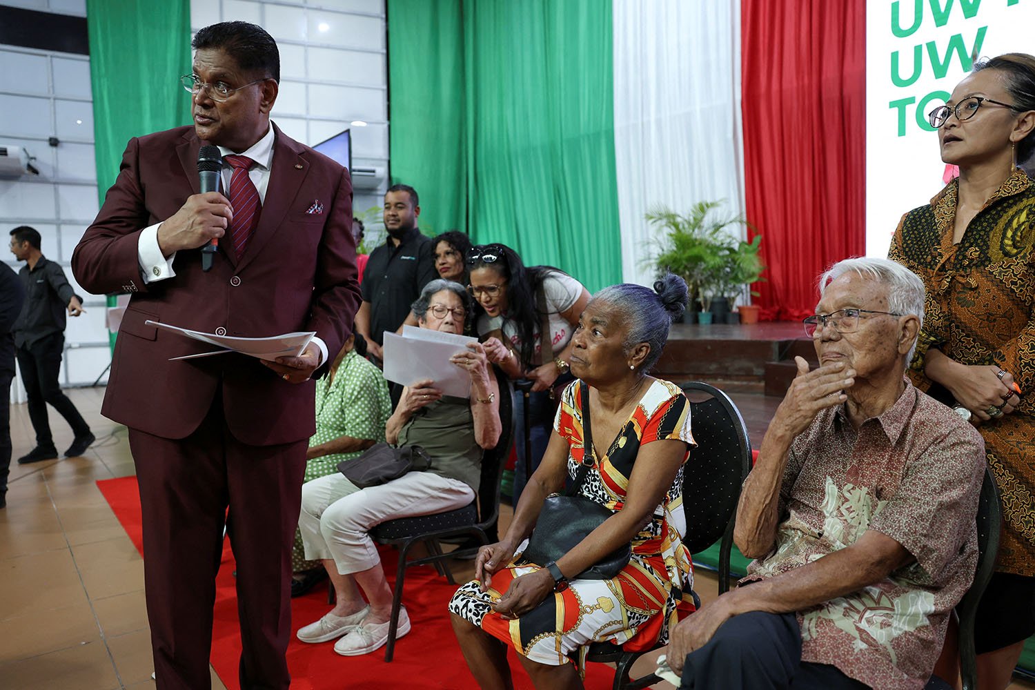 Santokhi in a suit holds a microphone and papers in his hand as he stands next to elderly seated people in a room draped in green, white and red.