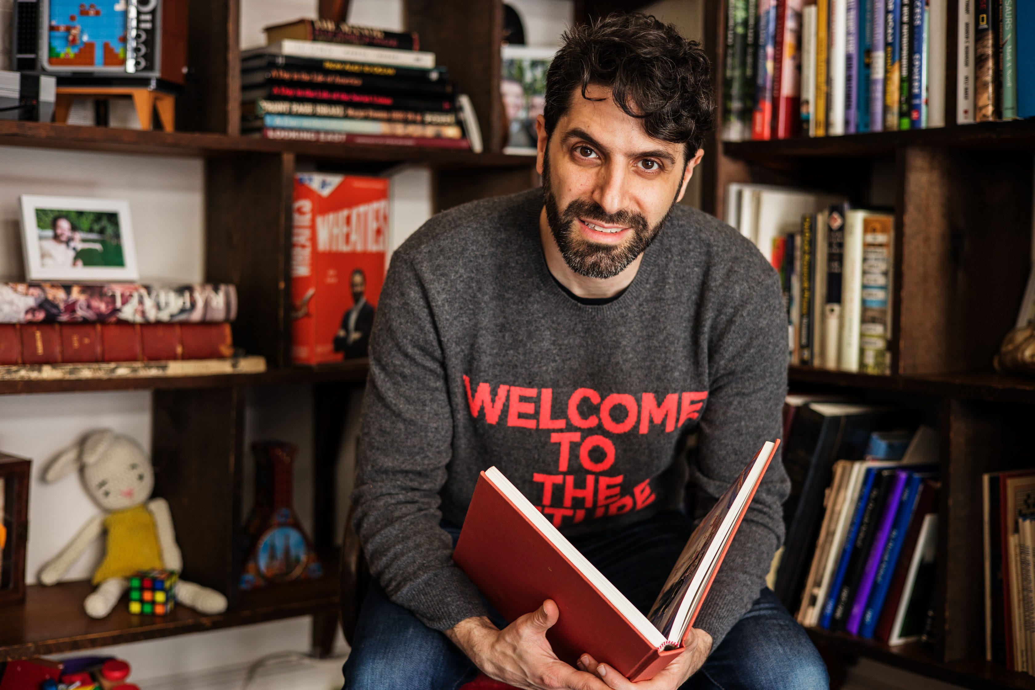 A man in a gray sweater sits in front of a book shelf, holding a red book open
