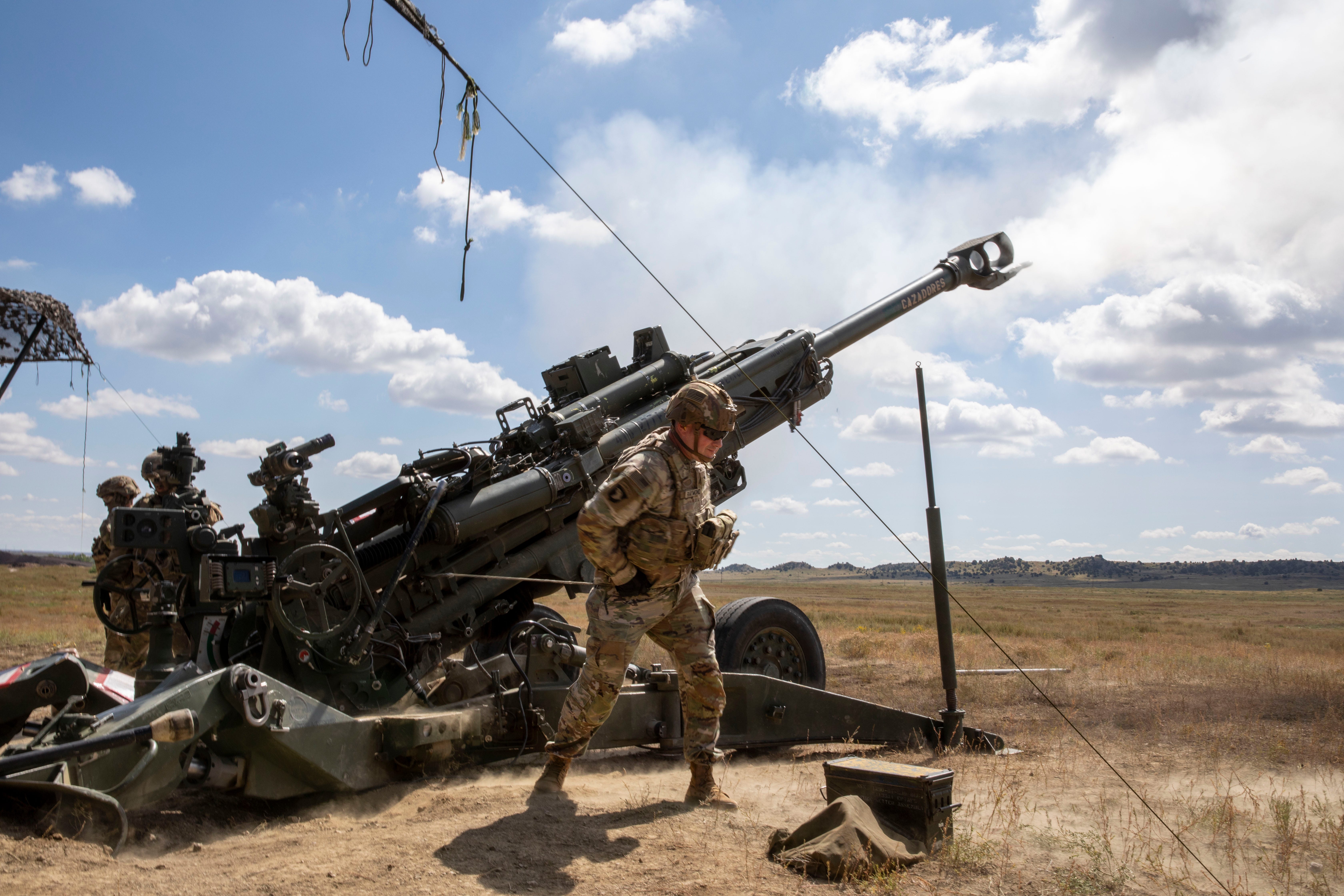 A soldier wearing camouflage stands in front of a black M777 Howitzer that's firing on a grassy field with a blue, cloudy sky in the background.
