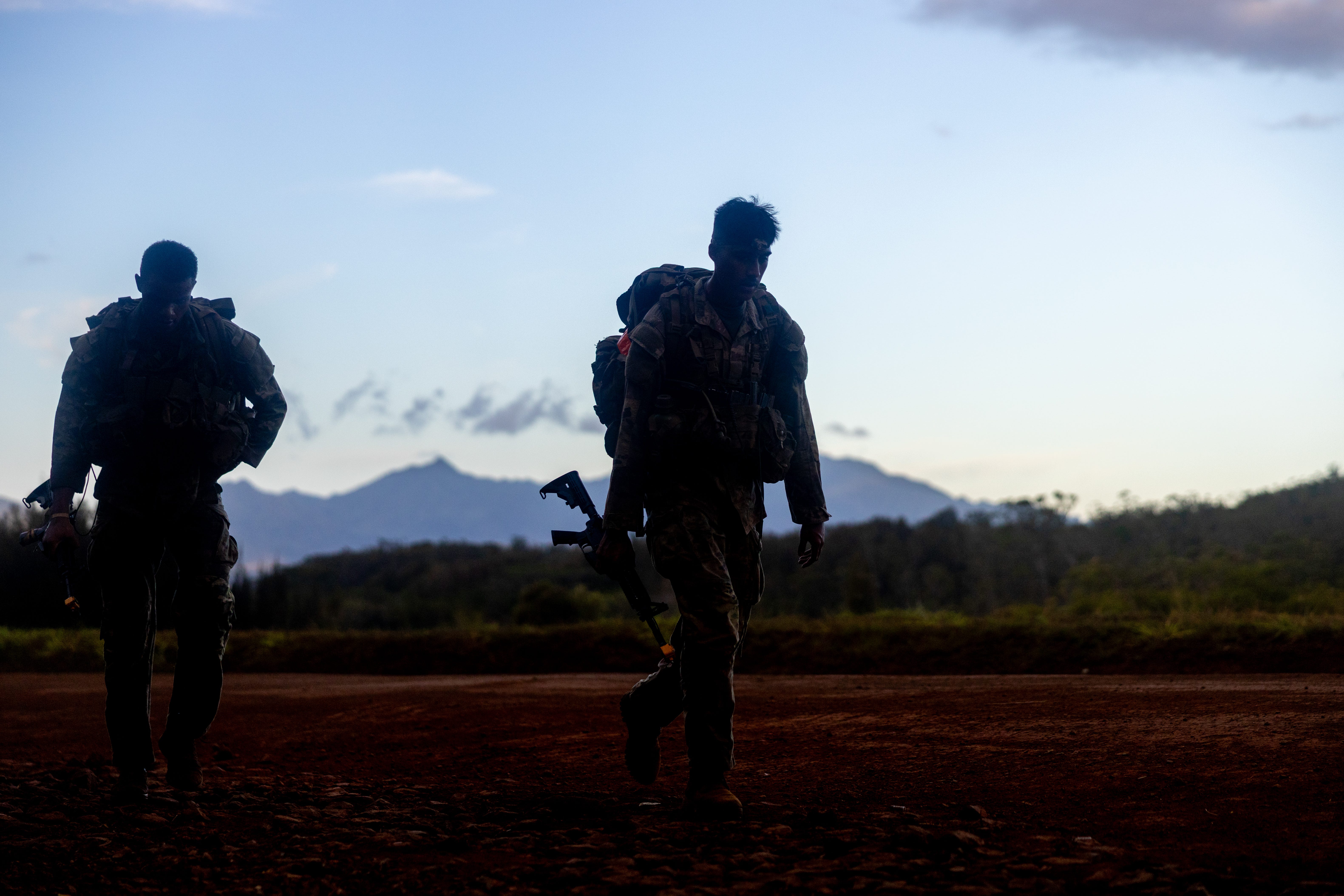 Two US Army soldiers walk on a red dirt road with lines of fresh and clouds in the background. The sky is blue.