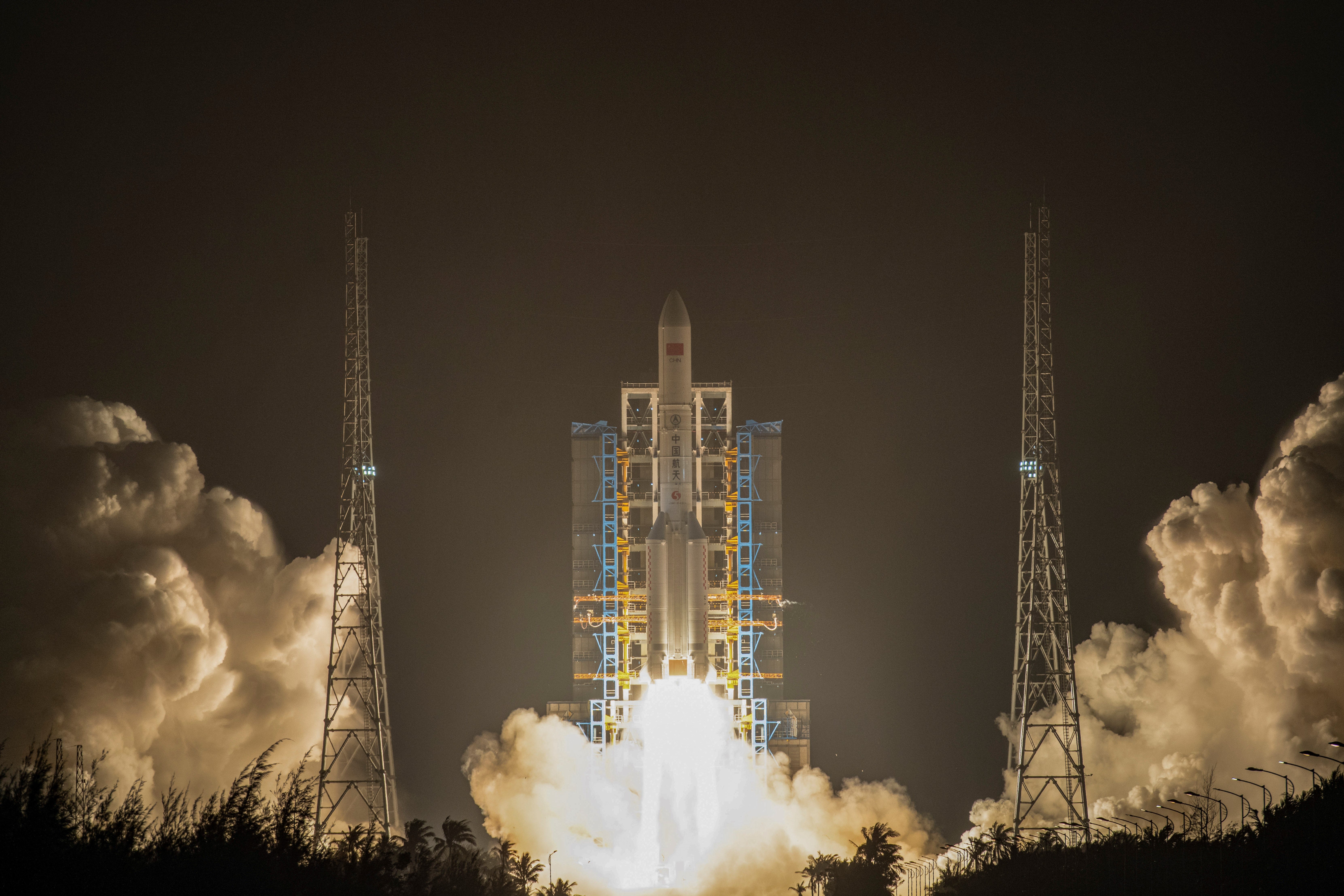 A Chinese rocket launches at night, with a row of trees at the bottom of the picture and two large plumes of smoke on either side of the launch.
