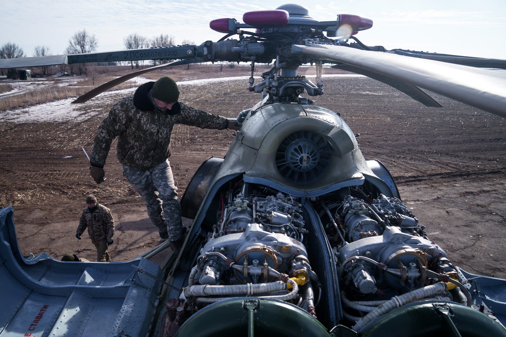 A man wearing camouflage gear and a beanie stands on top of a helicopter that is sat on a brown ground, with some of its internal systems exposed