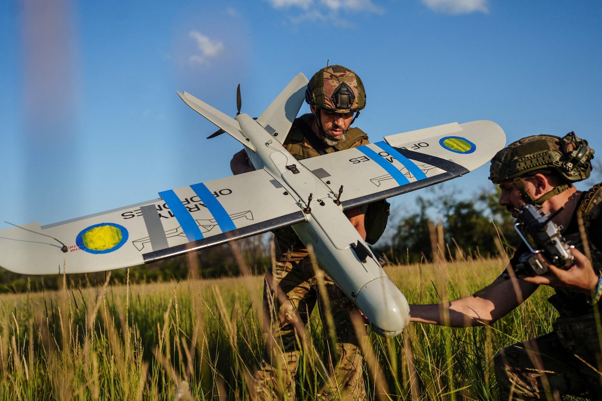 A man in camoflage gear and a helmet stands holding a large grey drone with blue tape on it, with another man assisting, in tall grass and under a blue sky