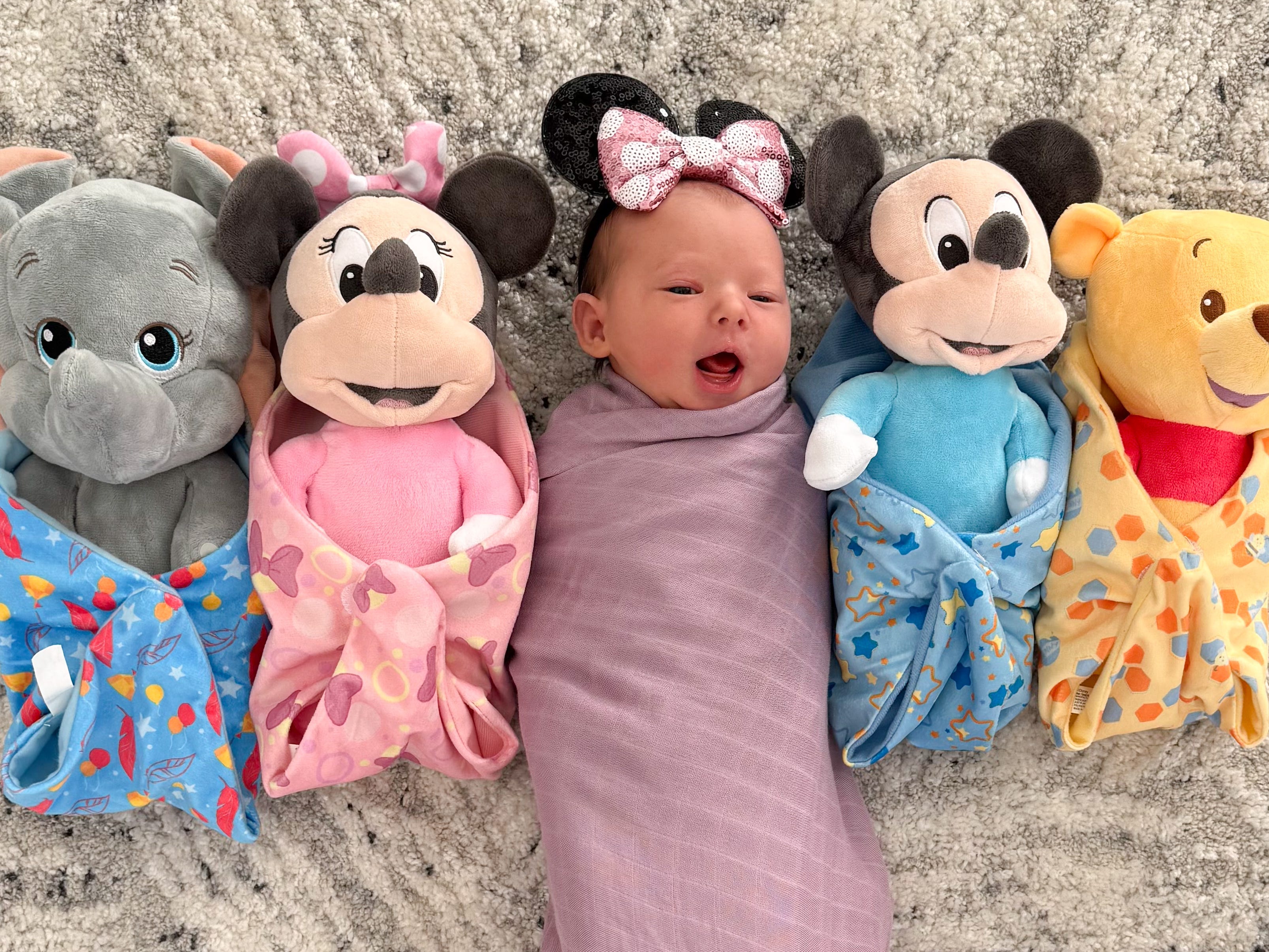 A newborn wearing Mickey Mouse ears lying next to some stuffed toys