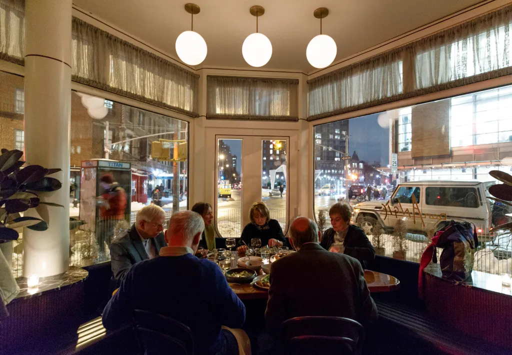 Diners seated at a table inside Don Angie restaurant with a view of a snowy Greenwich Village street at night.
