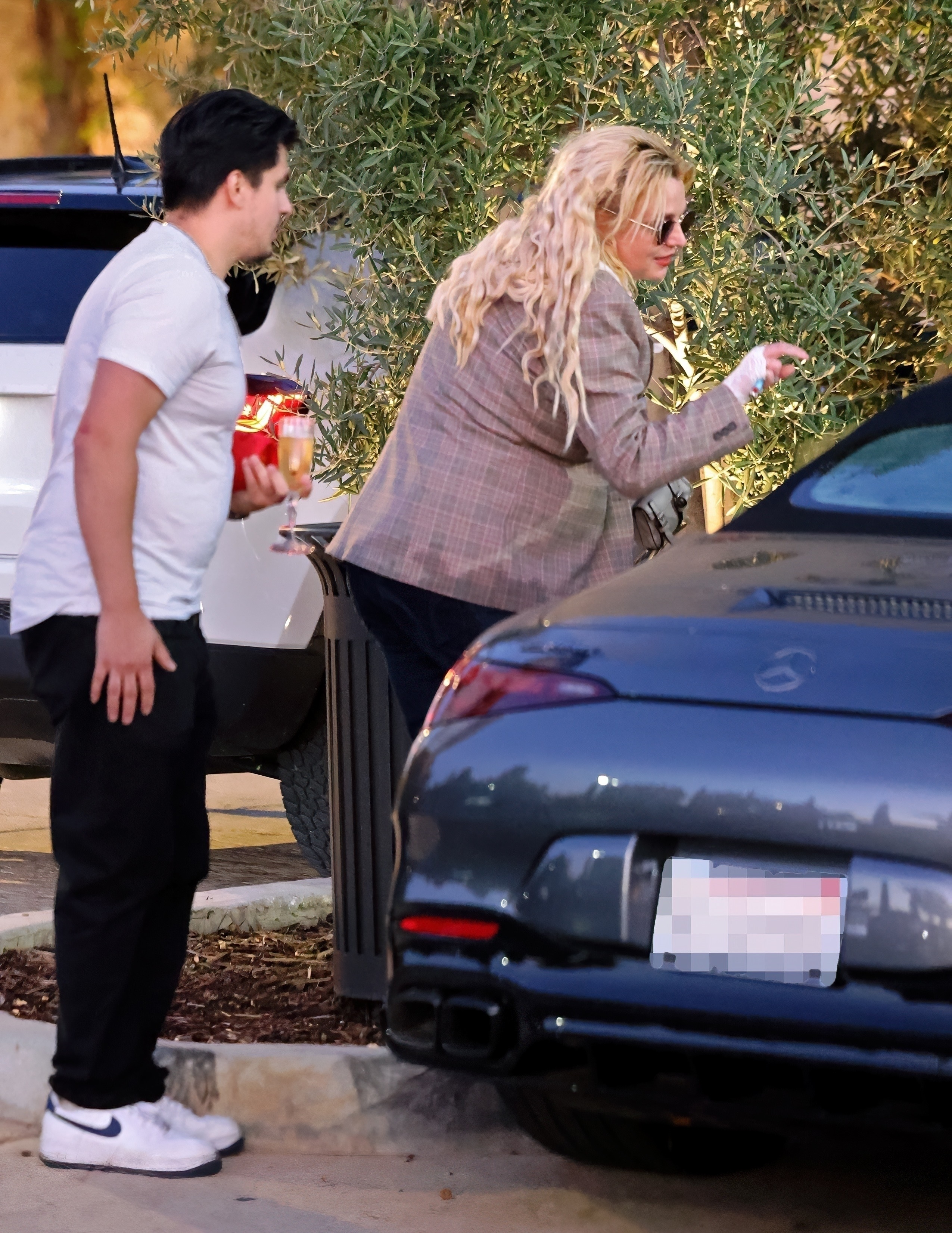 Britney Spears wearing mirrored sunglasses, a plaid blazer, and white lace gloves, standing next to a man holding a champagne flute, in front of a grey Mercedes car.