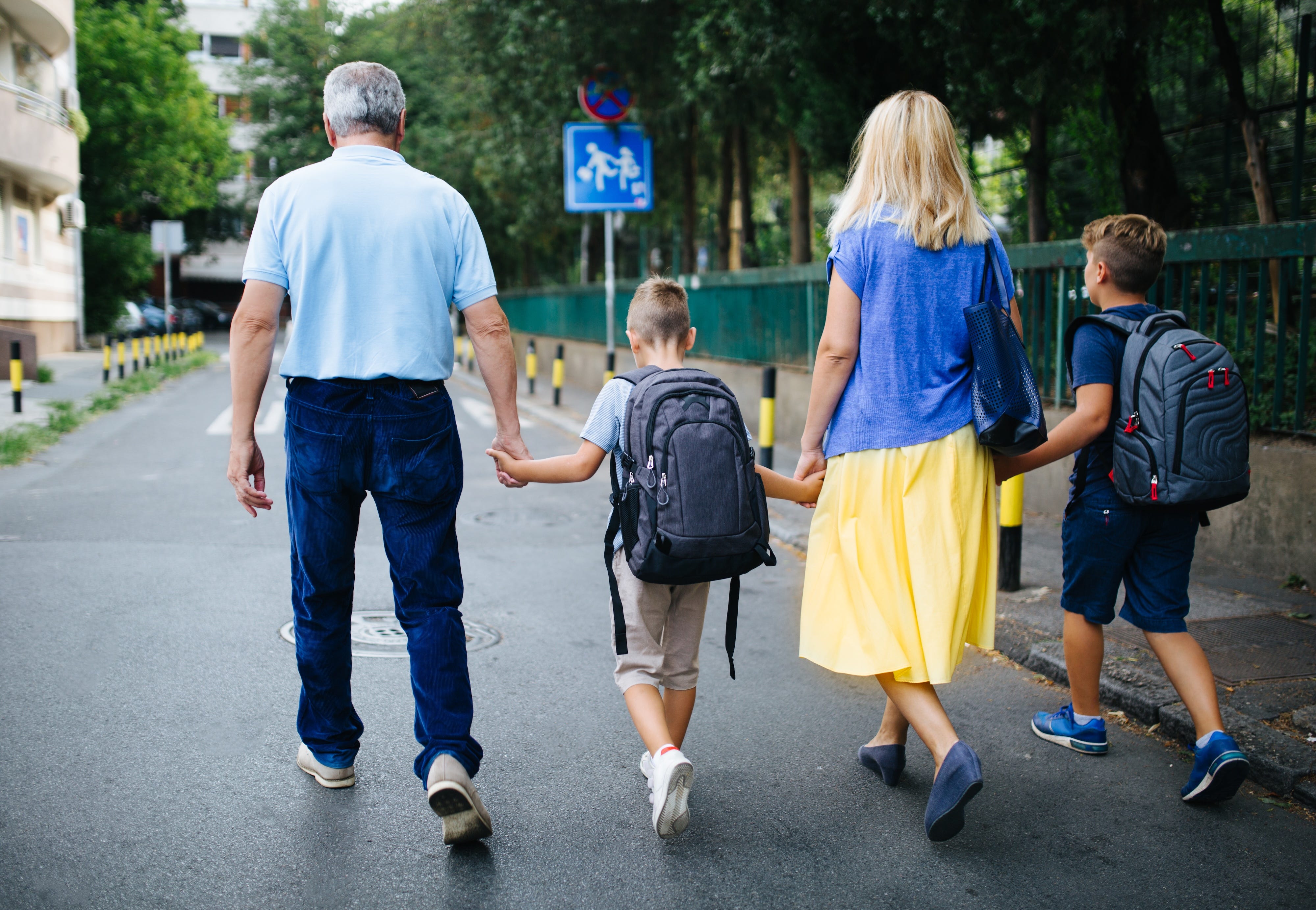 Grandparents walking down the street holding hands with their grandkids, who are wearing backpacks.