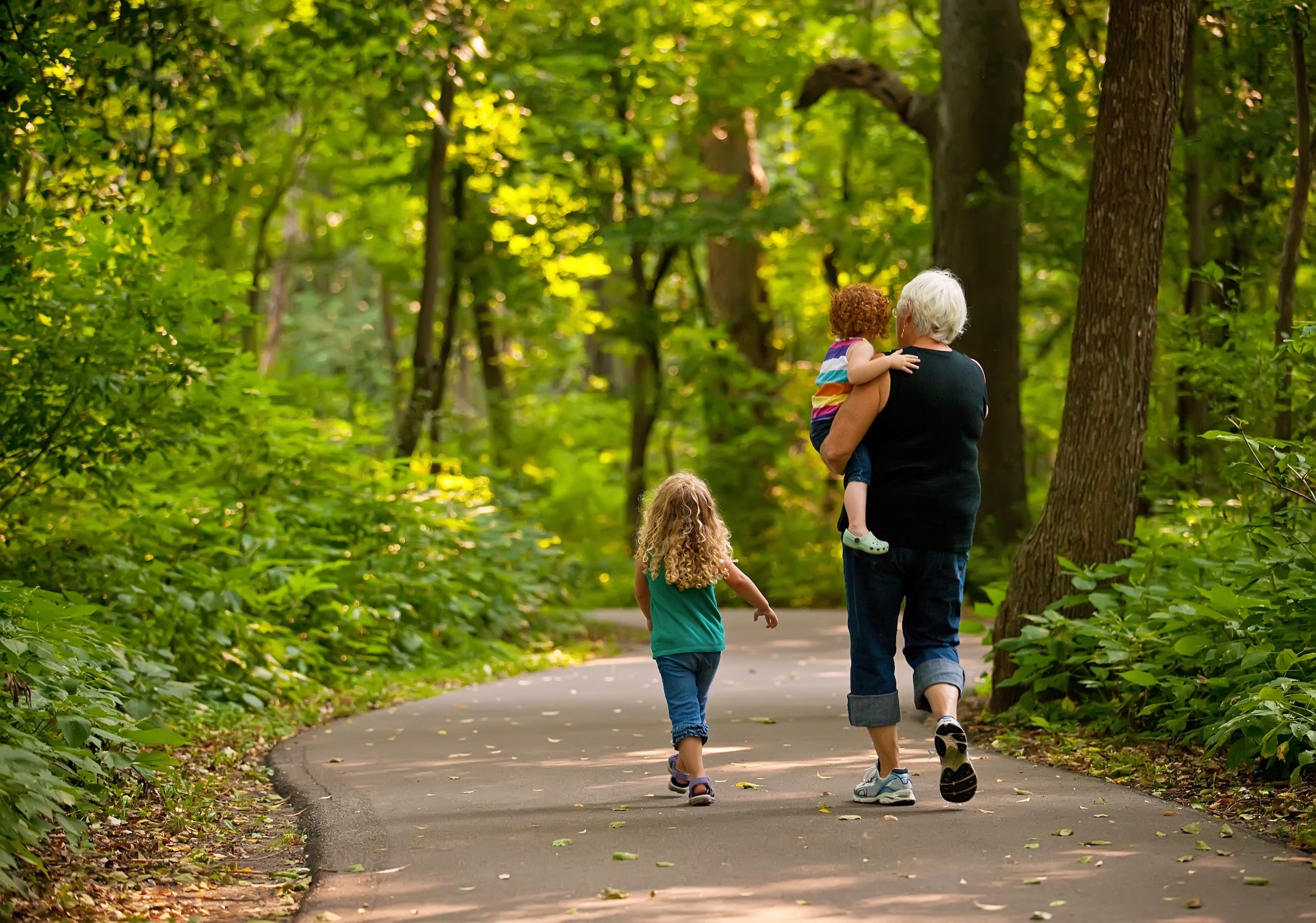 A grandmother walking down a path, holding one child while one child walks beside her.