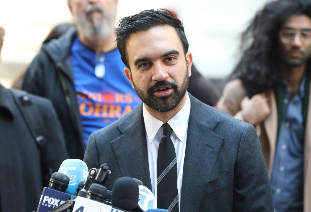 Zohran Mamdani speaking at a press conference surrounded by microphones.