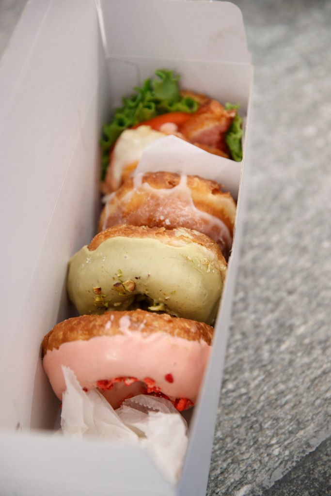 Donuts in a box with pink, green, and white icing, and one savory donut with lettuce and meat.