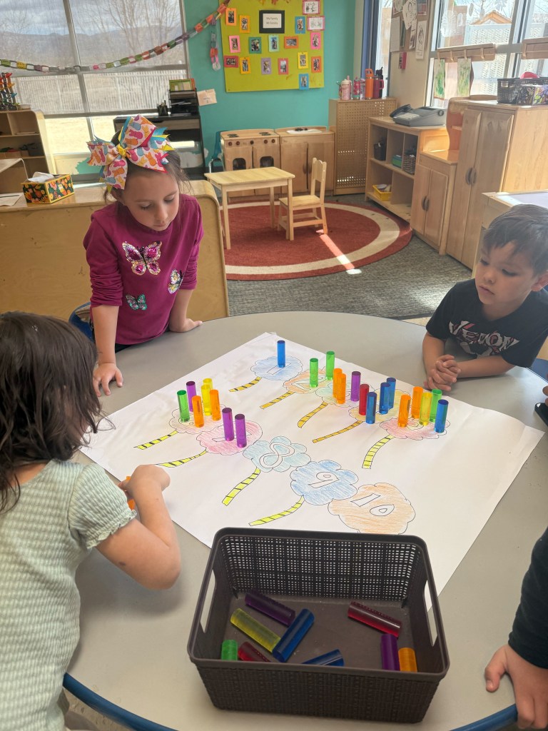 Three children playing with colorful cylinders and a large drawing of numbers on a table in a classroom.