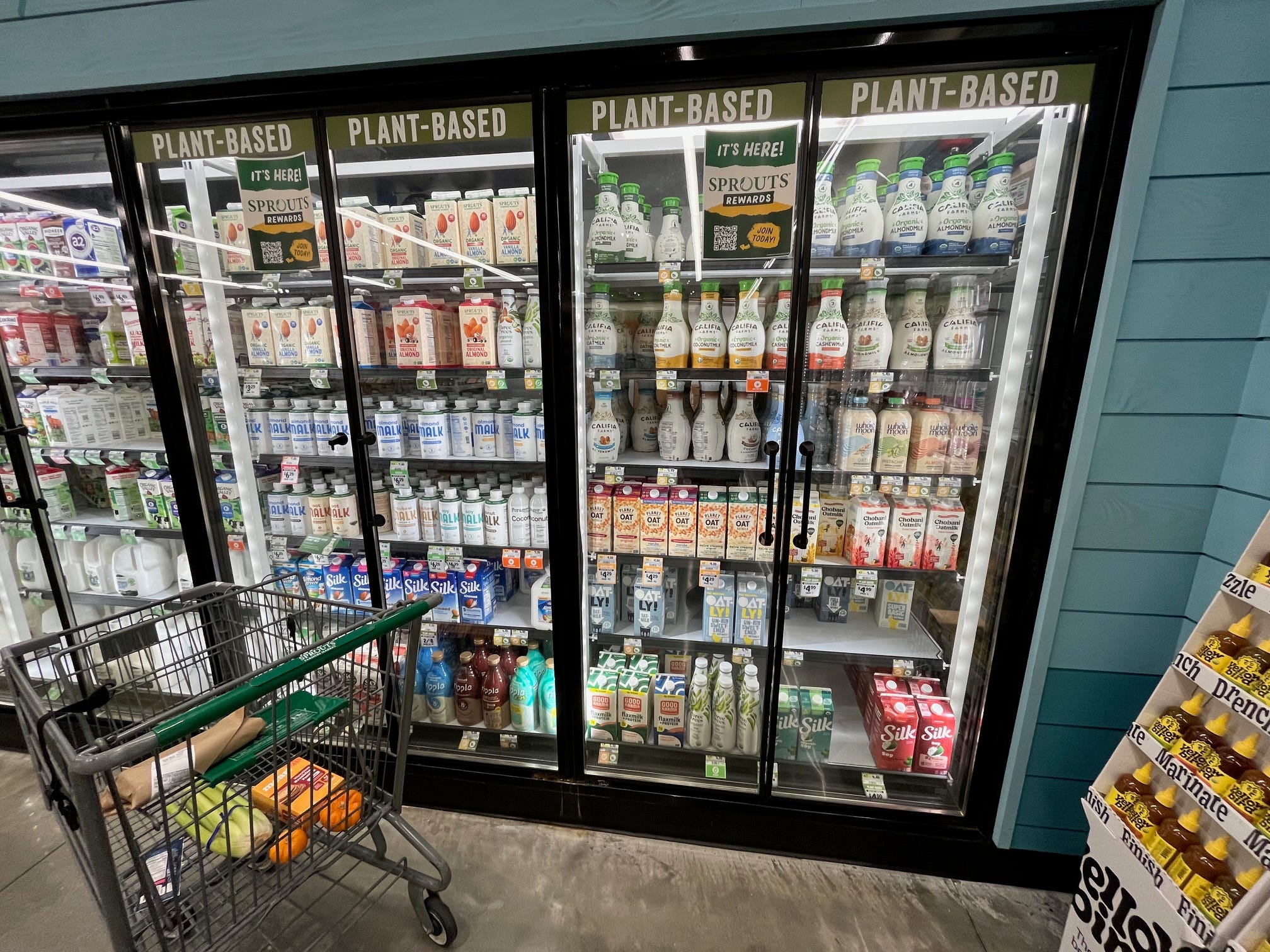 Plant-based milks sit in a cooler case with signs above reading