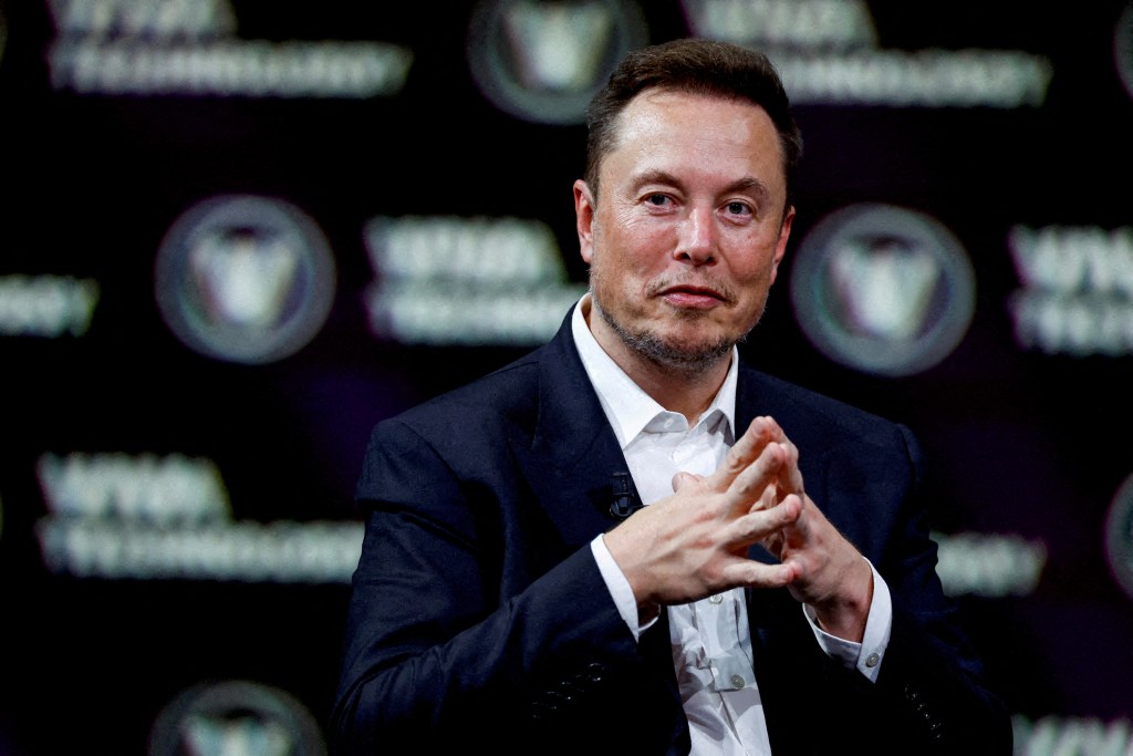 Elon Musk with his hands clasped, wearing a dark suit jacket and a white collared shirt, against a dark blurred background.