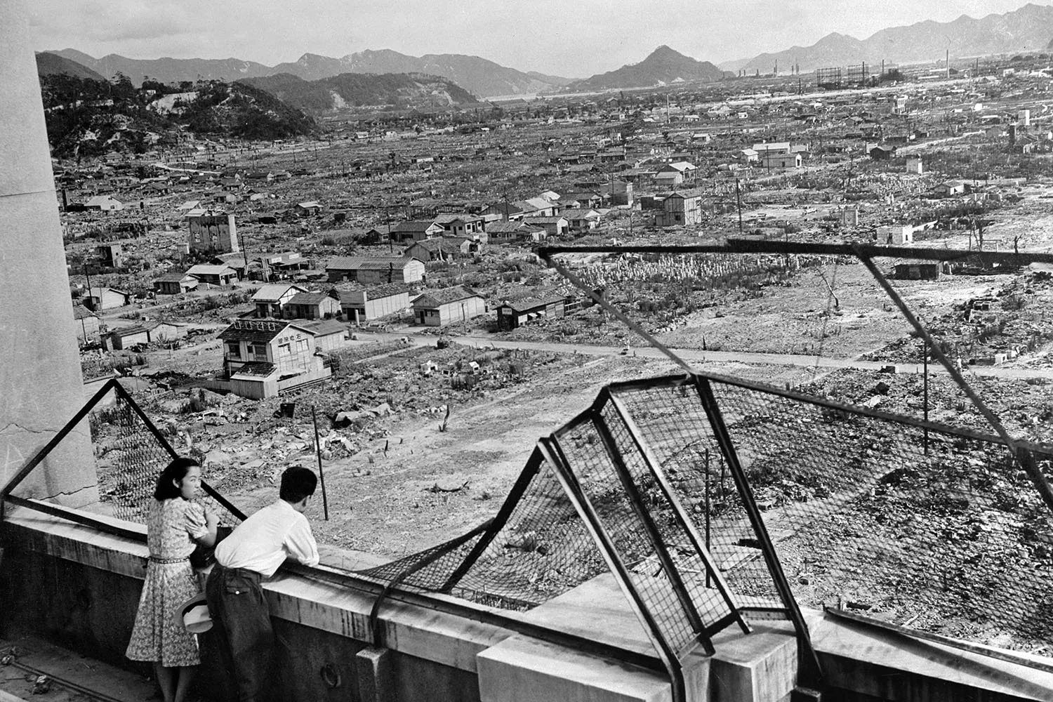 Two people stand on a ledge overlooking the grid of a city with the debris of demolished buildings stretching to the horizon.